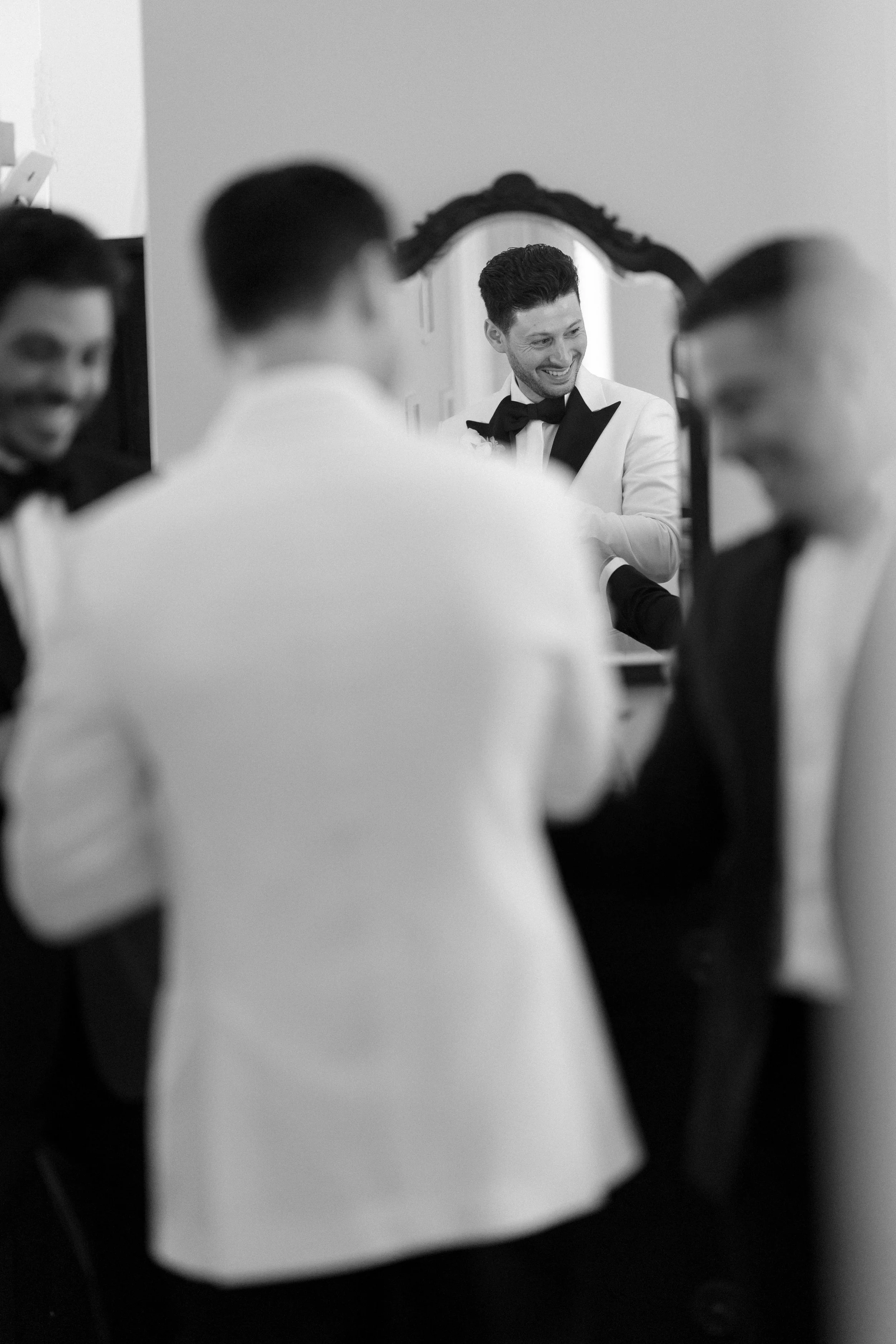 A man in a tuxedo with a bow tie is smiling while looking at himself in a mirror, surrounded by other men also dressed in tuxedos, getting ready for a formal event.