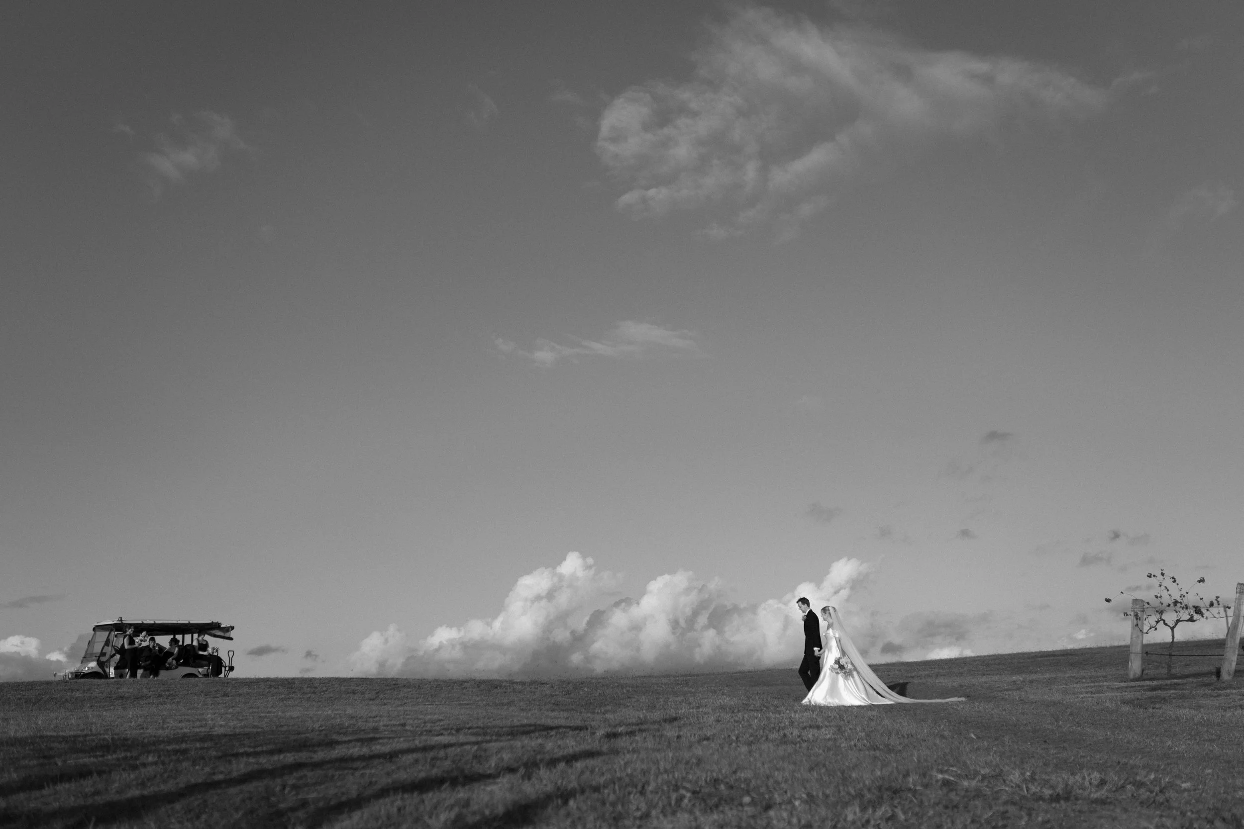 A black-and-white photo of a couple walking hand in hand on a grassy field. The woman wears a long dress with a train, and the man is dressed in a suit. In the background, a golf cart with people sitting inside is visible under a partly cloudy sky.