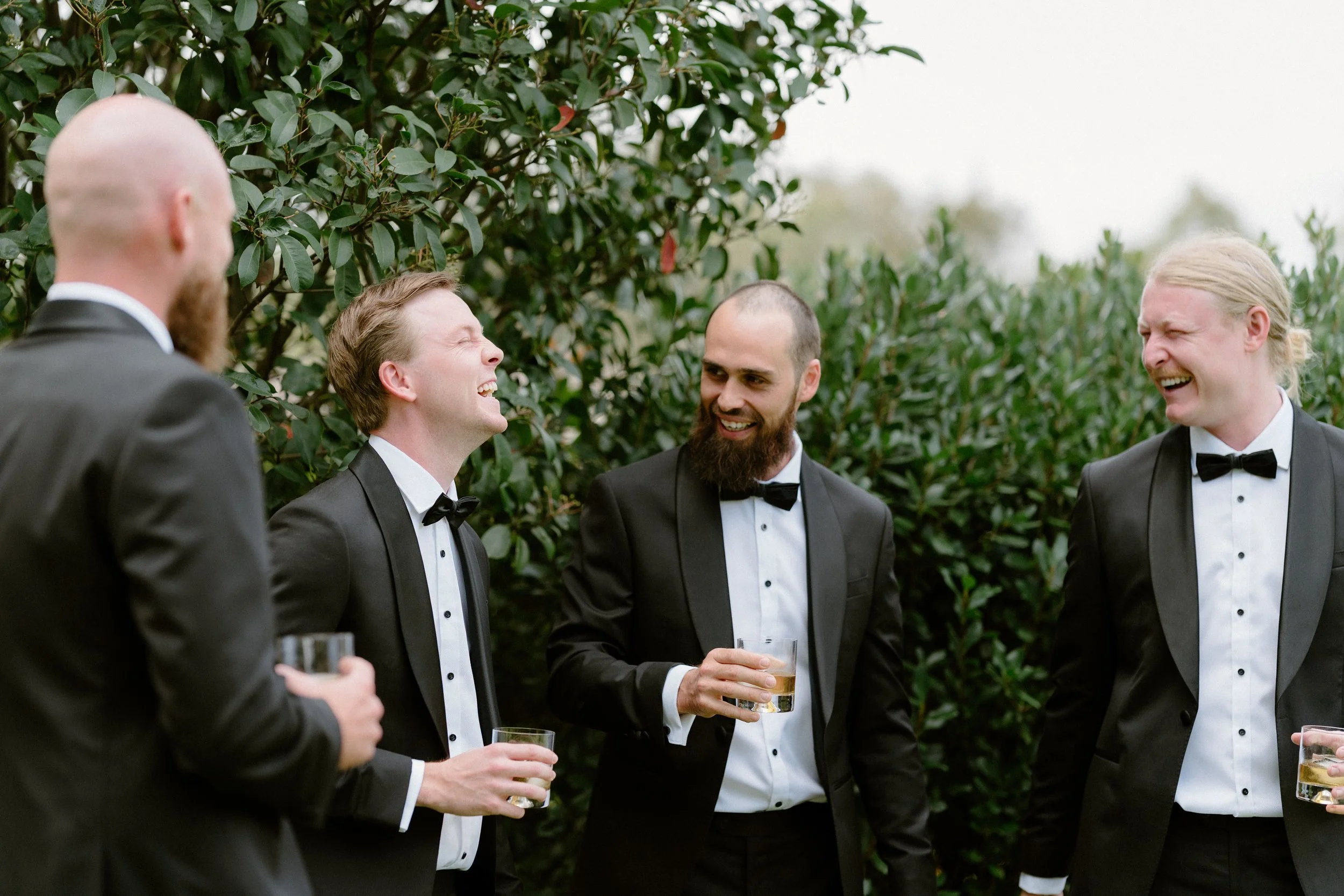 Group of men in tuxedos laughing and talking outdoors, holding drinks, with greenery in the background.