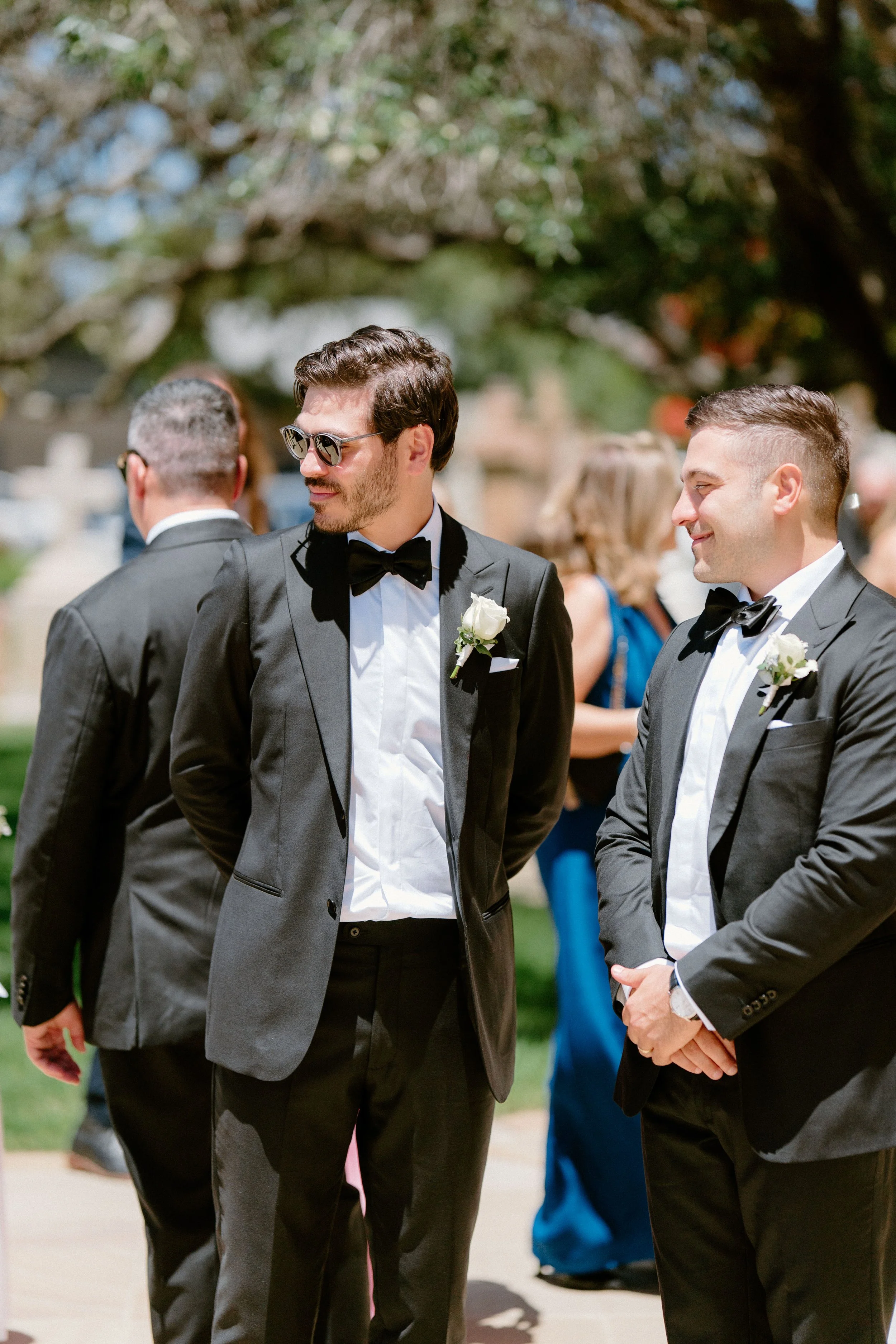 Two men dressed in black tuxedos with bow ties and white boutonnières, standing outdoors at a wedding or formal event, smiling and talking with other guests in the background.