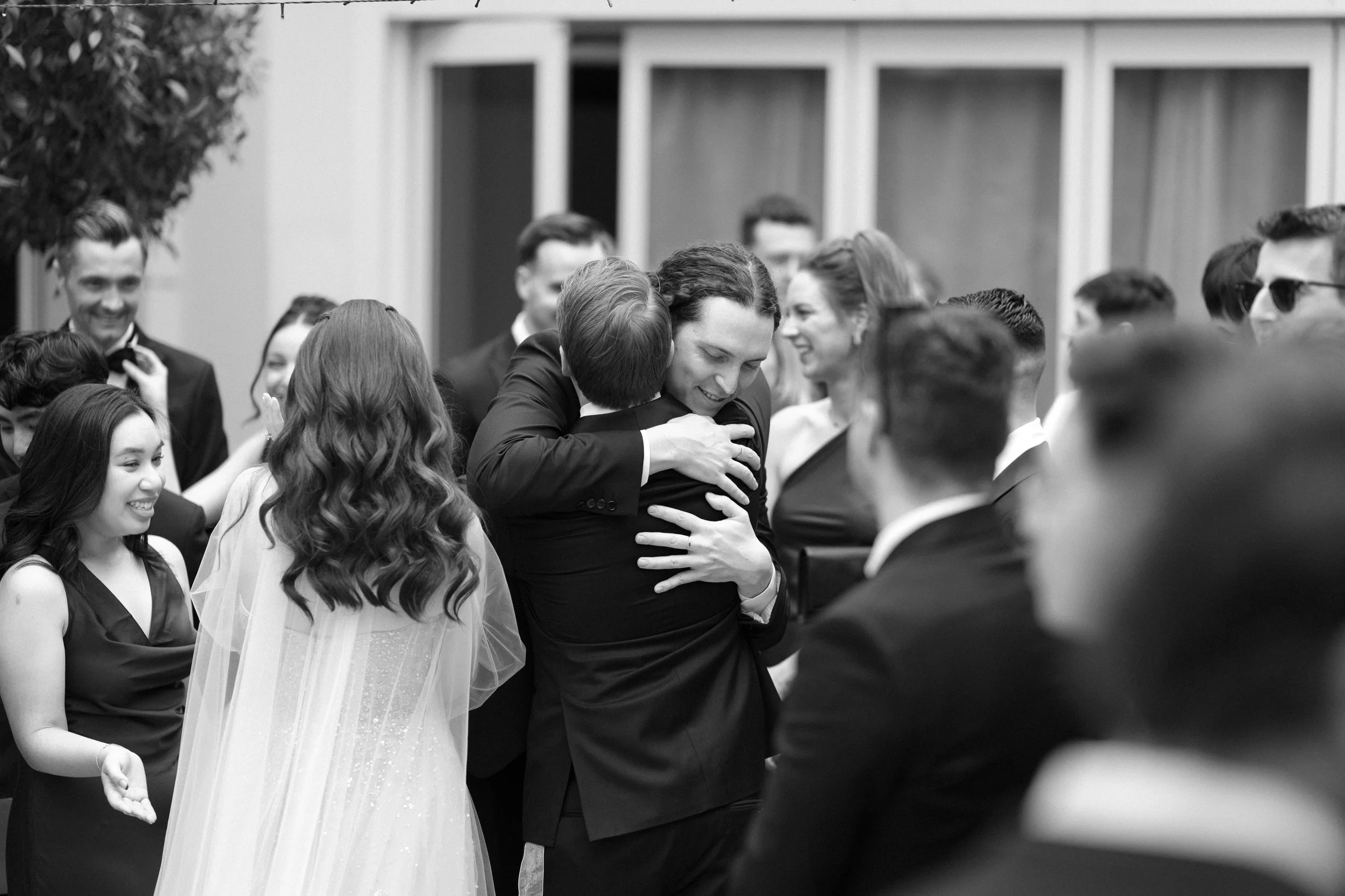 Black and white photo of people at a wedding, with a woman in a wedding dress hugging a man in a suit while others look on and smile.
