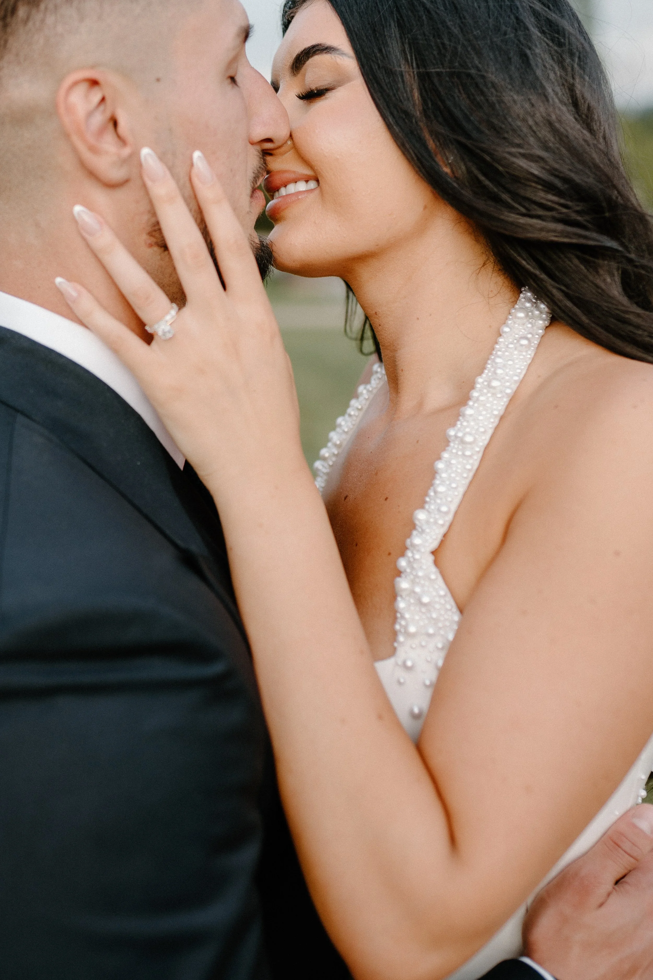 A bride and groom sharing a kiss outdoors, with the bride holding the groom's face and showing a wedding ring.