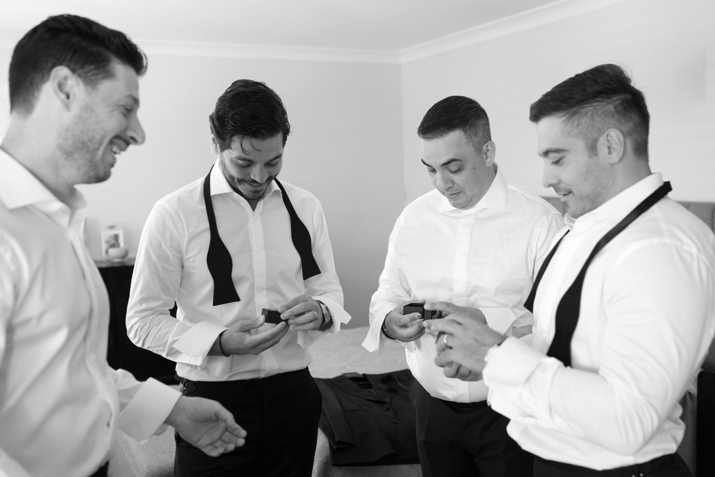 Four men in white shirts and light-colored pants, some with black ties, are standing in a room, looking at small boxes in their hands, smiling and engaging with each other.