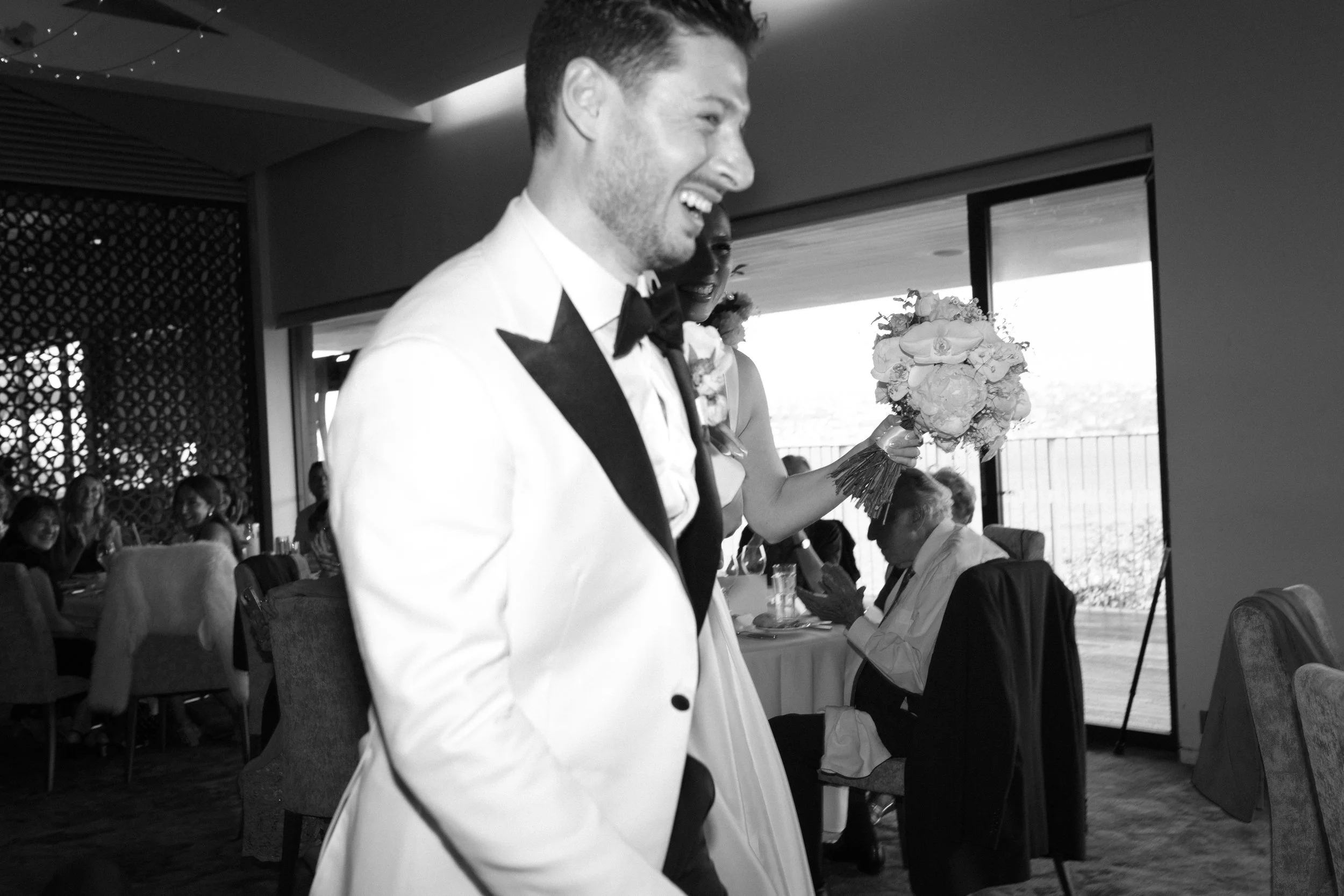 A man in a tuxedo with a black bow tie, smiling and looking to the side during a celebration. A woman holding a bouquet behind him and other guests are visible seated in a decorated dining area with large windows.