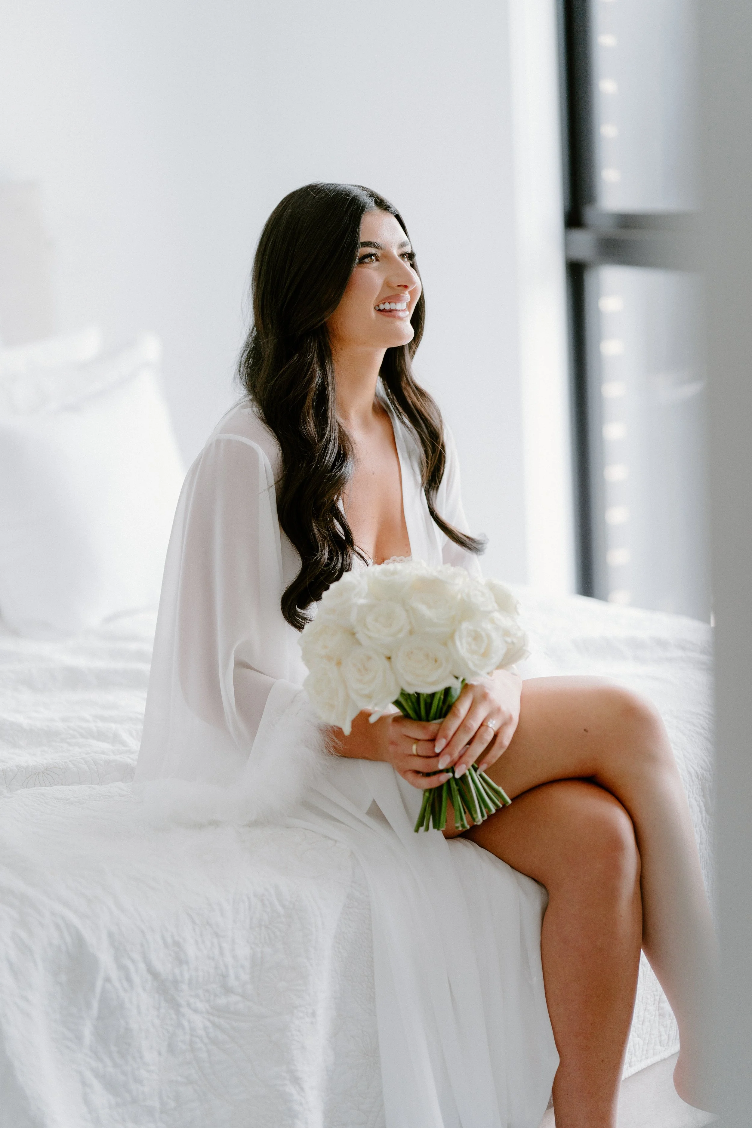 A woman sitting on a bed, holding a bouquet of white roses, smiling and looking out a window.