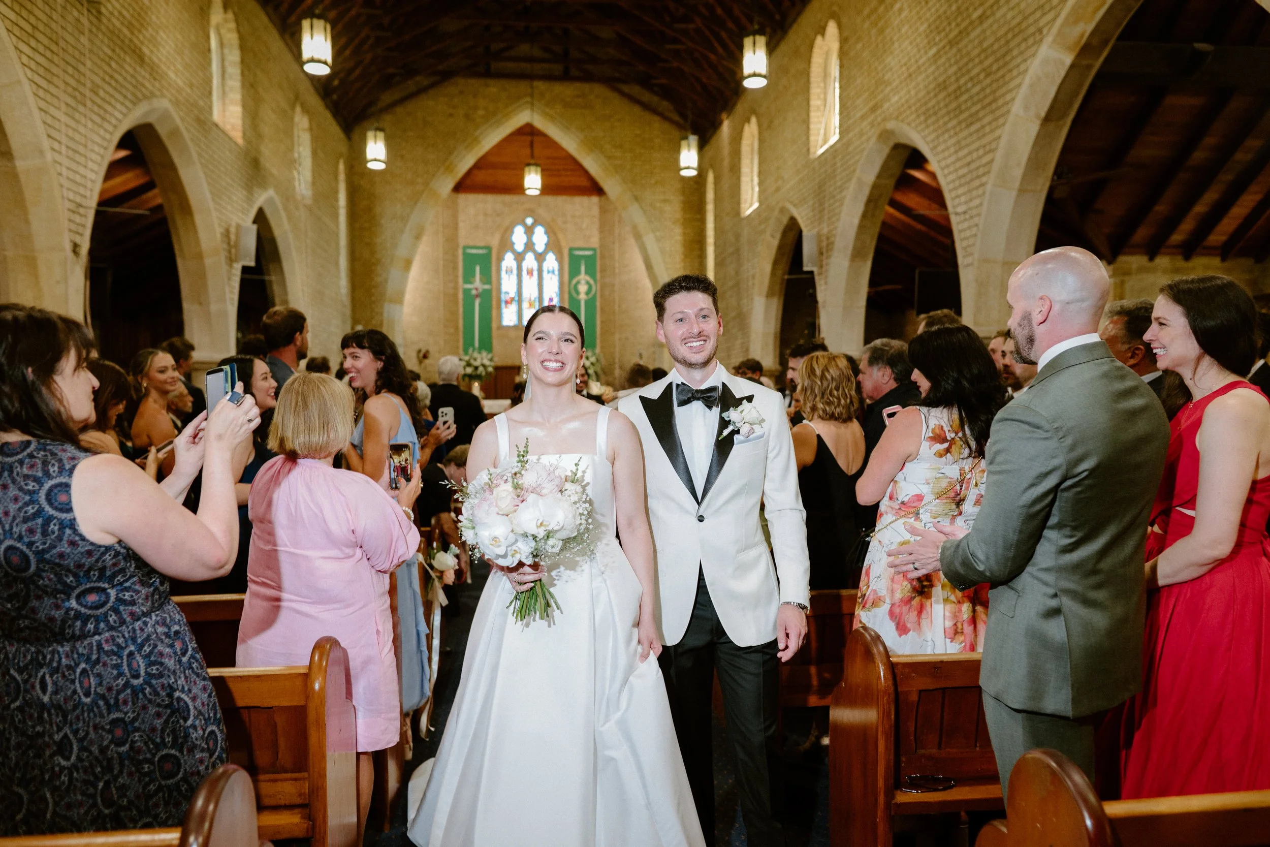 A bride and groom walking down the aisle of a church, smiling, with guests on either side taking photos and clapping. The church interior has high arched ceilings, stained glass windows, and hanging lights.