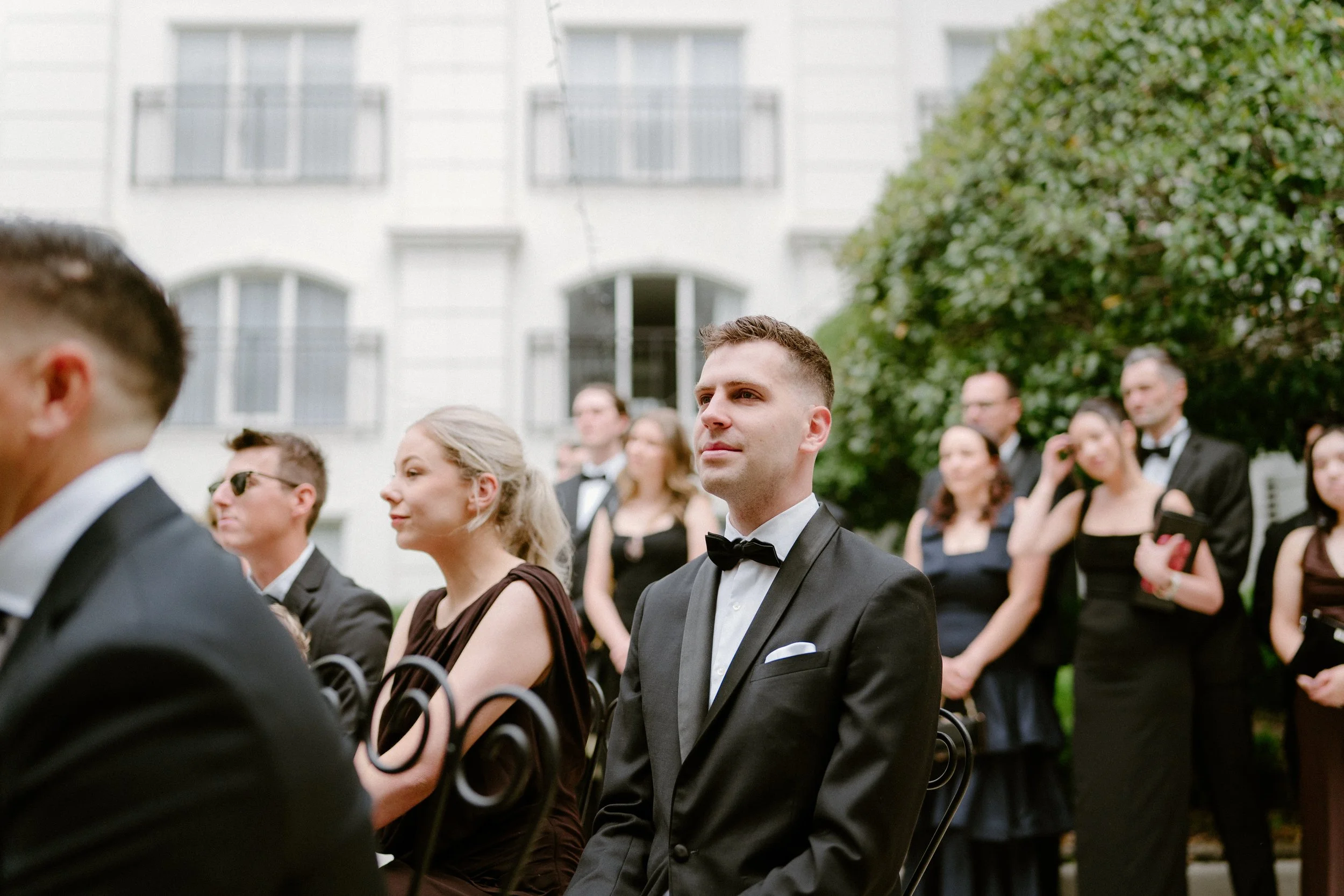 People attending an outdoor formal event, some in tuxedos and elegant dresses, seated and listening attentively in front of a white building and greenery.