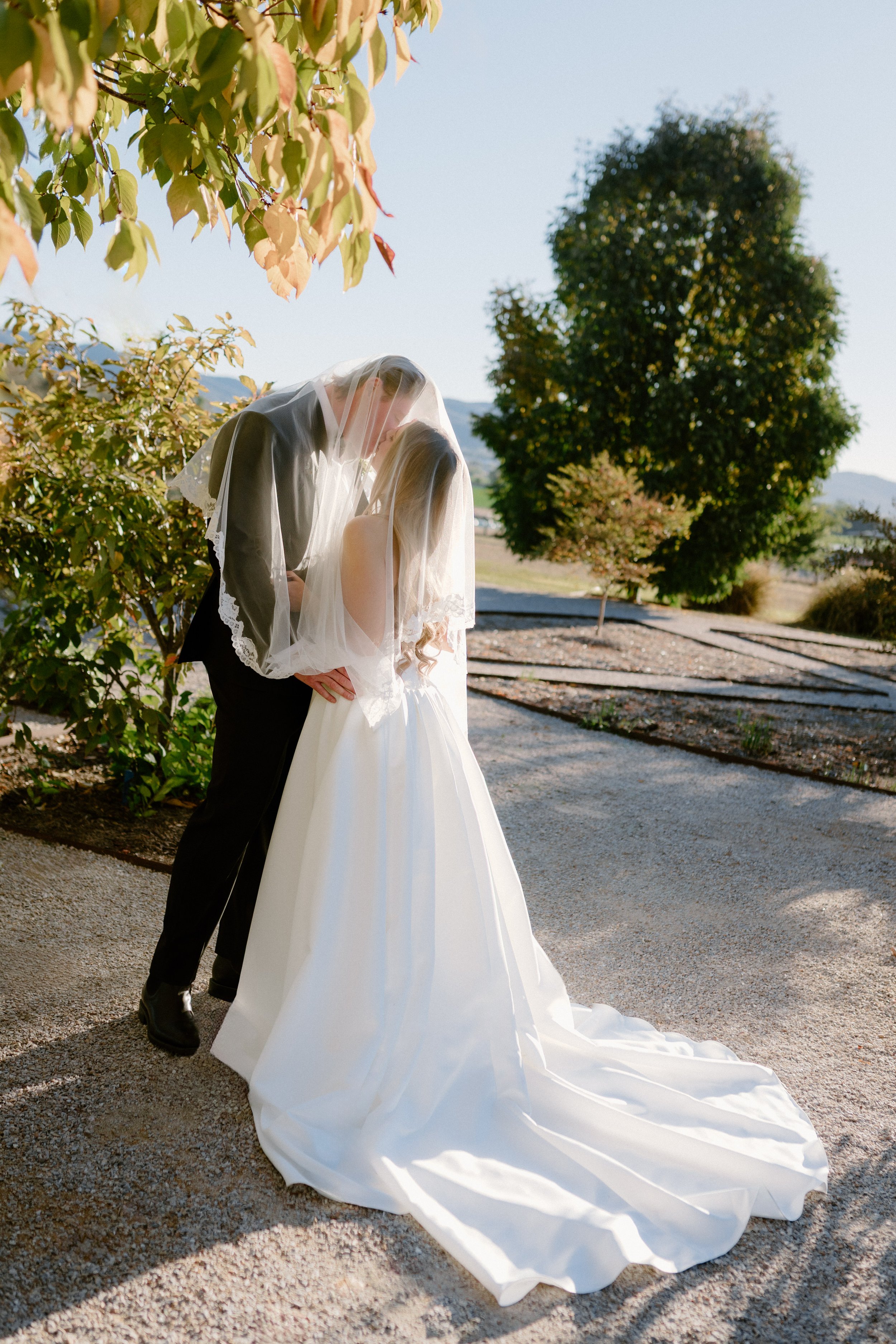 A bride and groom sharing a kiss outdoors, with the bride wearing a long white wedding gown and veil, and the groom in a black suit. They stand on a gravel path with trees and mountains in the background.