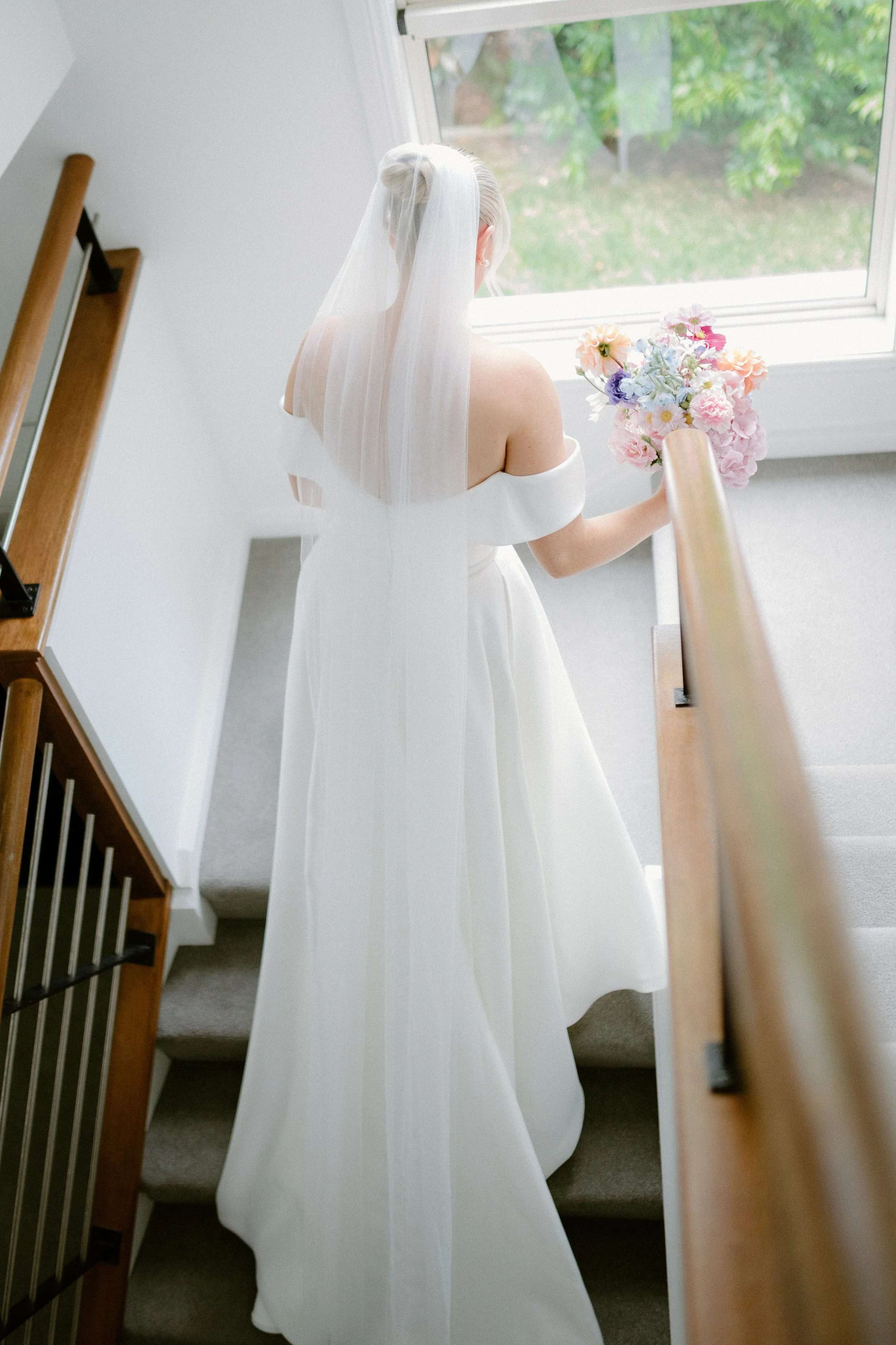 Bride in a white wedding dress with a veil holding a bouquet of pastel-colored flowers, standing at the top of a staircase near a large window.