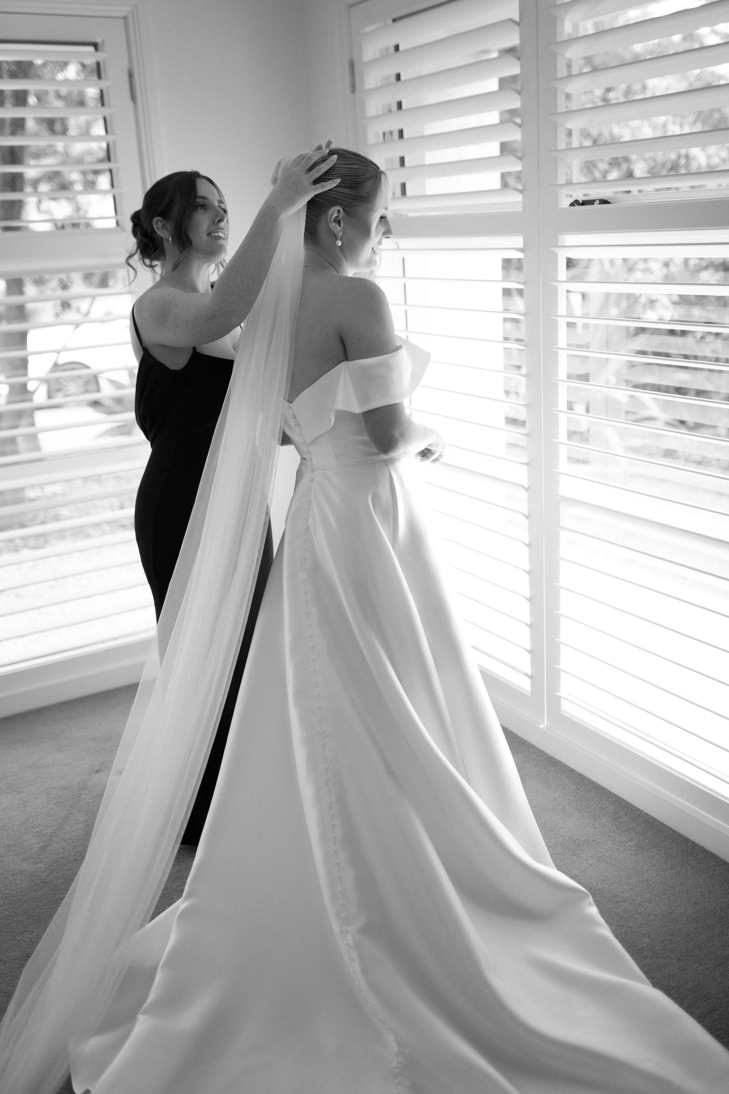 A bride wearing a white wedding dress stands by window shutters, looking outside with a smile. A woman, possibly a bridesmaid or family member, adjusts the bride's veil from behind. Bimbadgen Palmers Lane Wedding, Hunter Valley