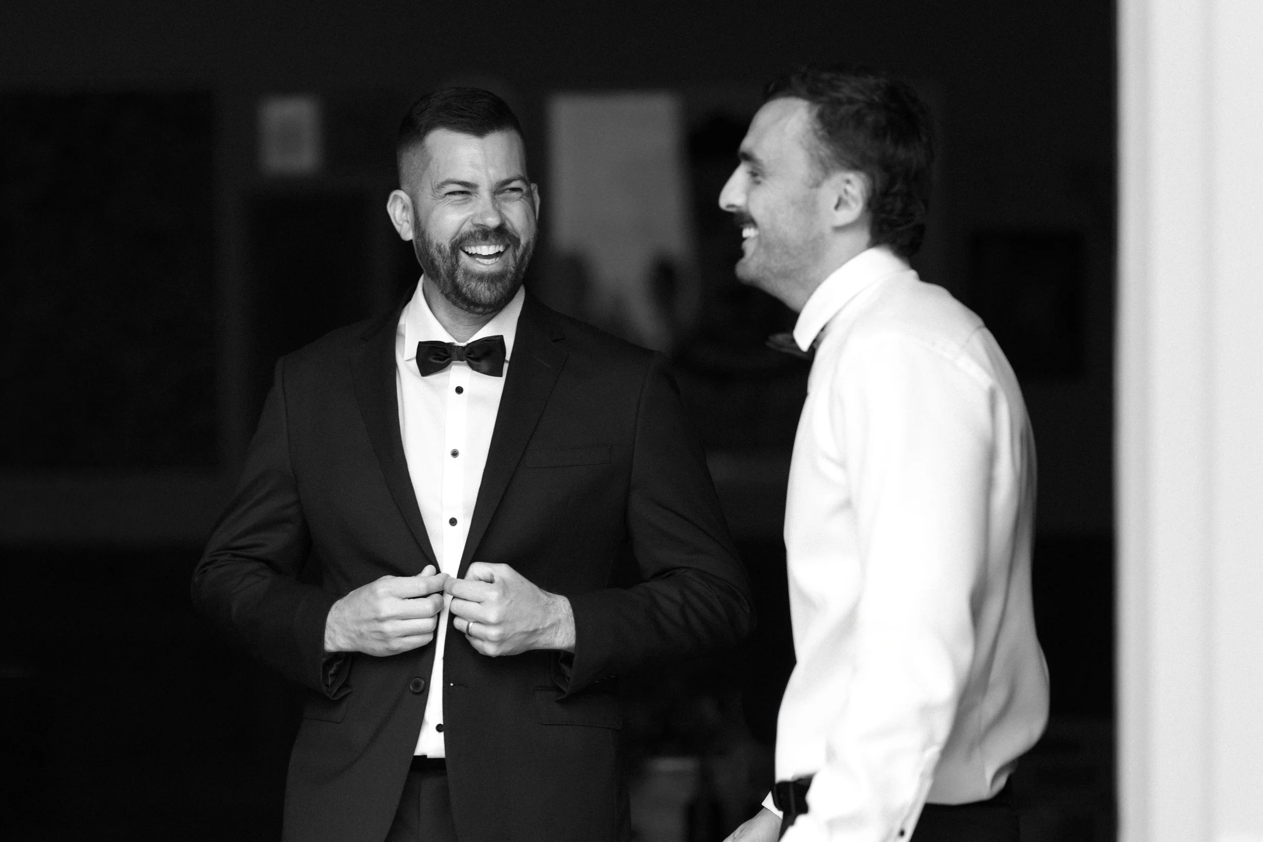 Two men dressed in formal tuxedos, laughing and enjoying a conversation.