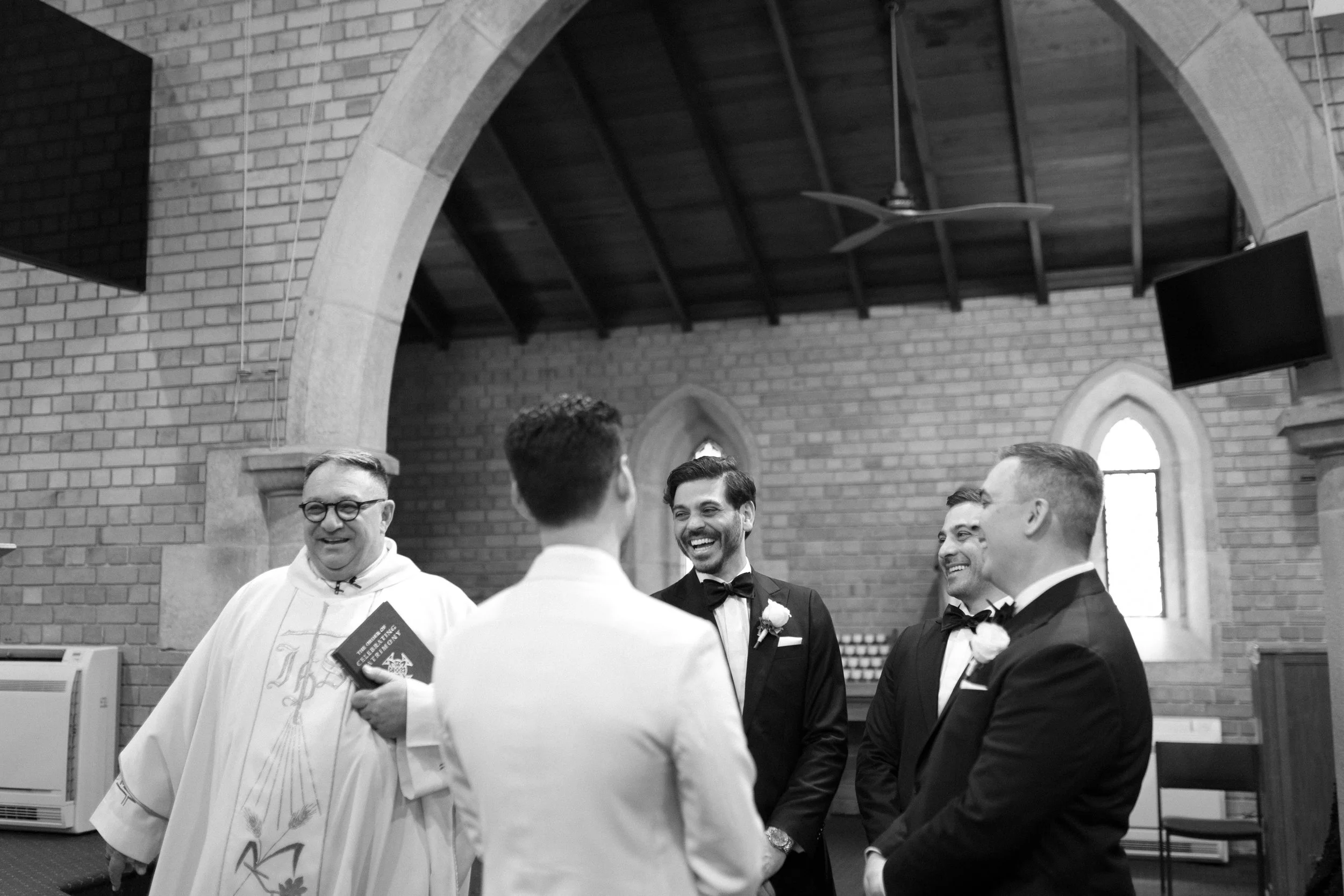 A wedding ceremony in a church with five men, including the groom and three groomsmen in tuxedos, and a priest in religious robes, all smiling and engaging in a moment of joy.