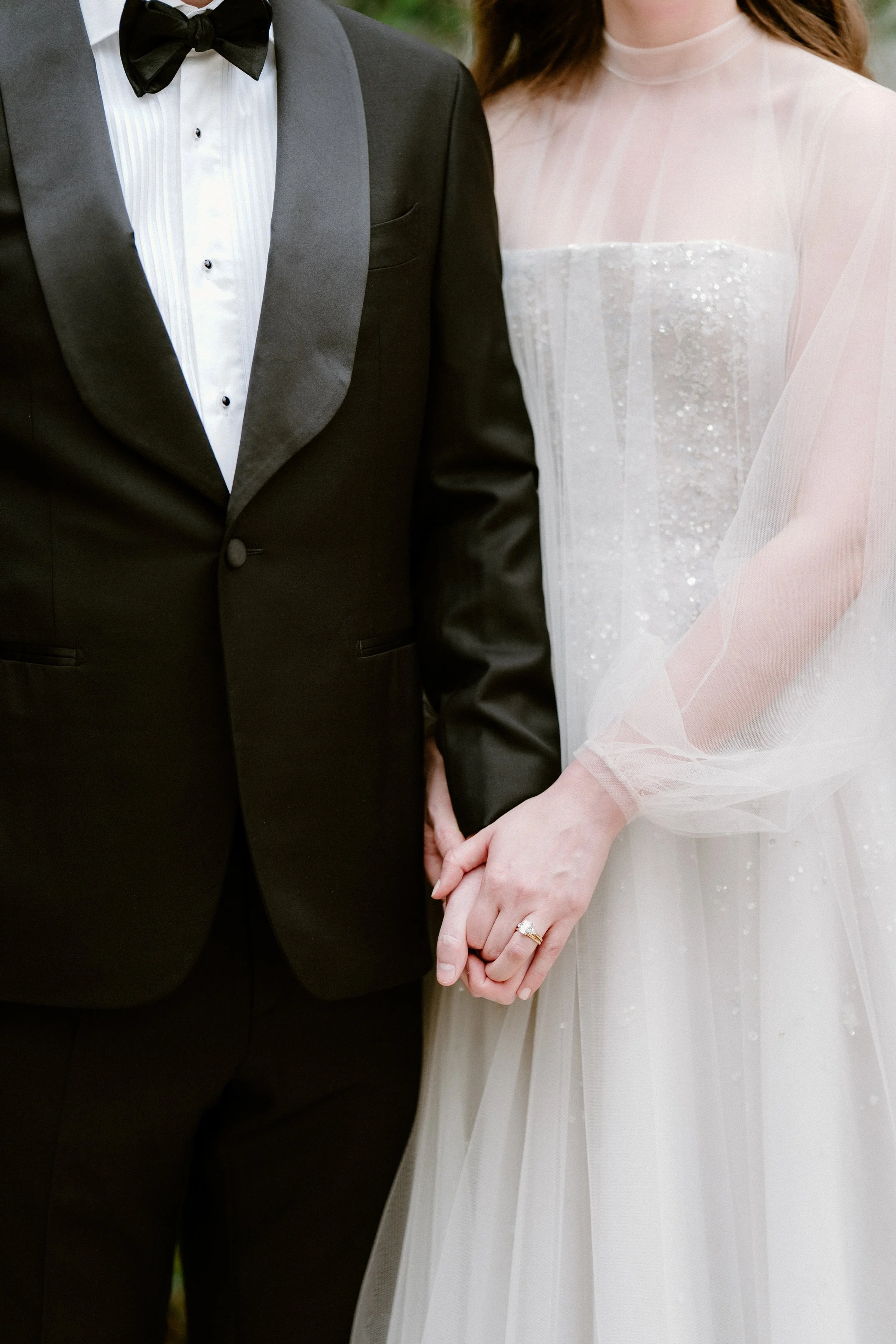 Close-up of a bride and groom holding hands at their wedding, focusing on the bride's wedding ring and the couple's formal wedding attire.