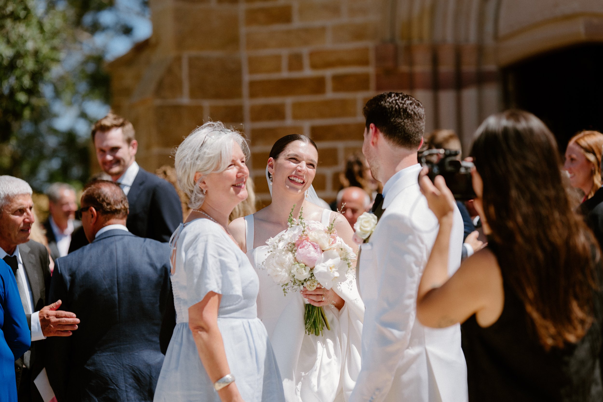 A bride and groom exchange vows outside a brick building, surrounded by smiling guests, with the bride holding a bouquet of white and pink flowers.