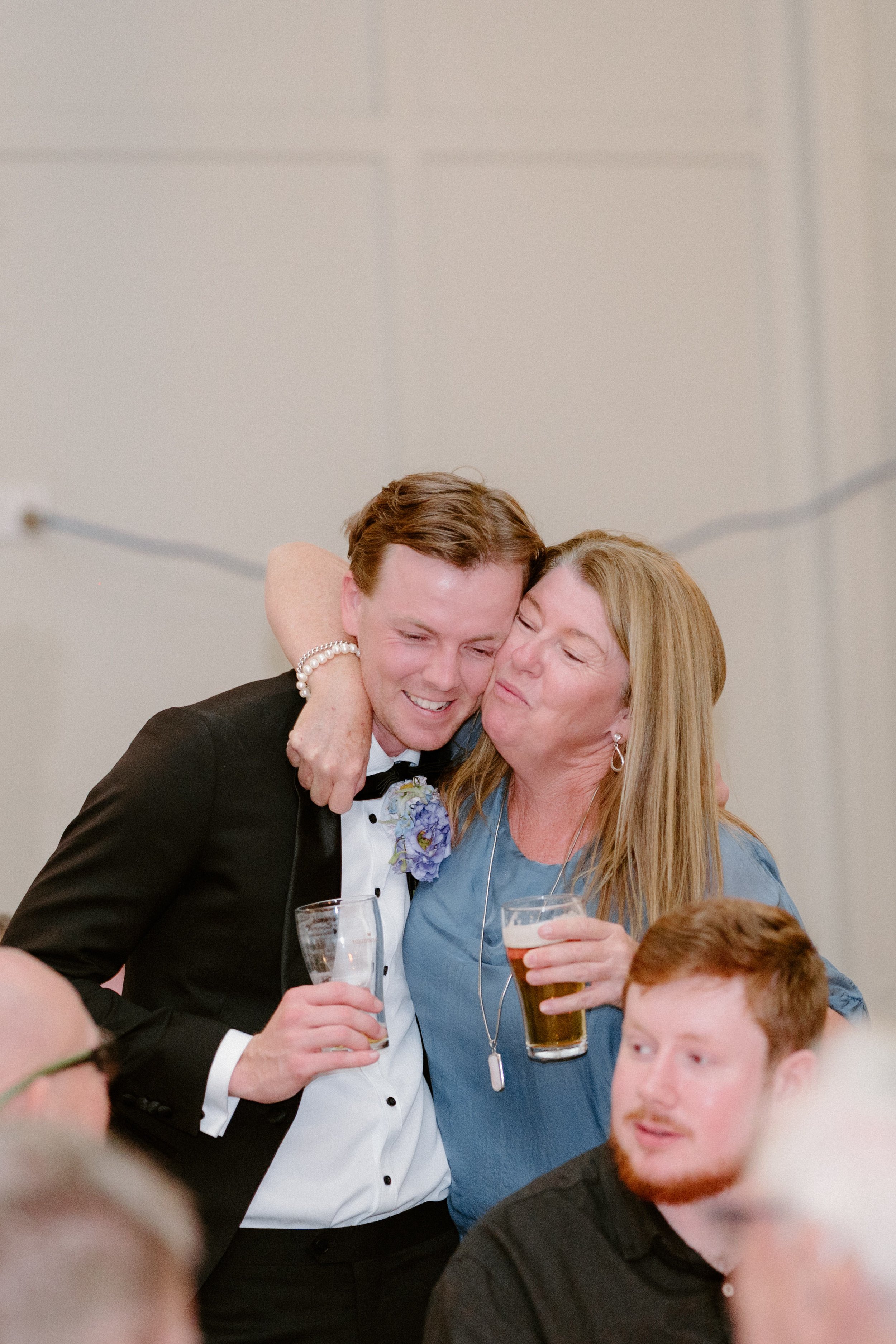 A young man in a tuxedo and an older woman in a blue dress sharing a hug at a celebration, with the young man holding a glass of champagne and the woman holding a glass of beer.