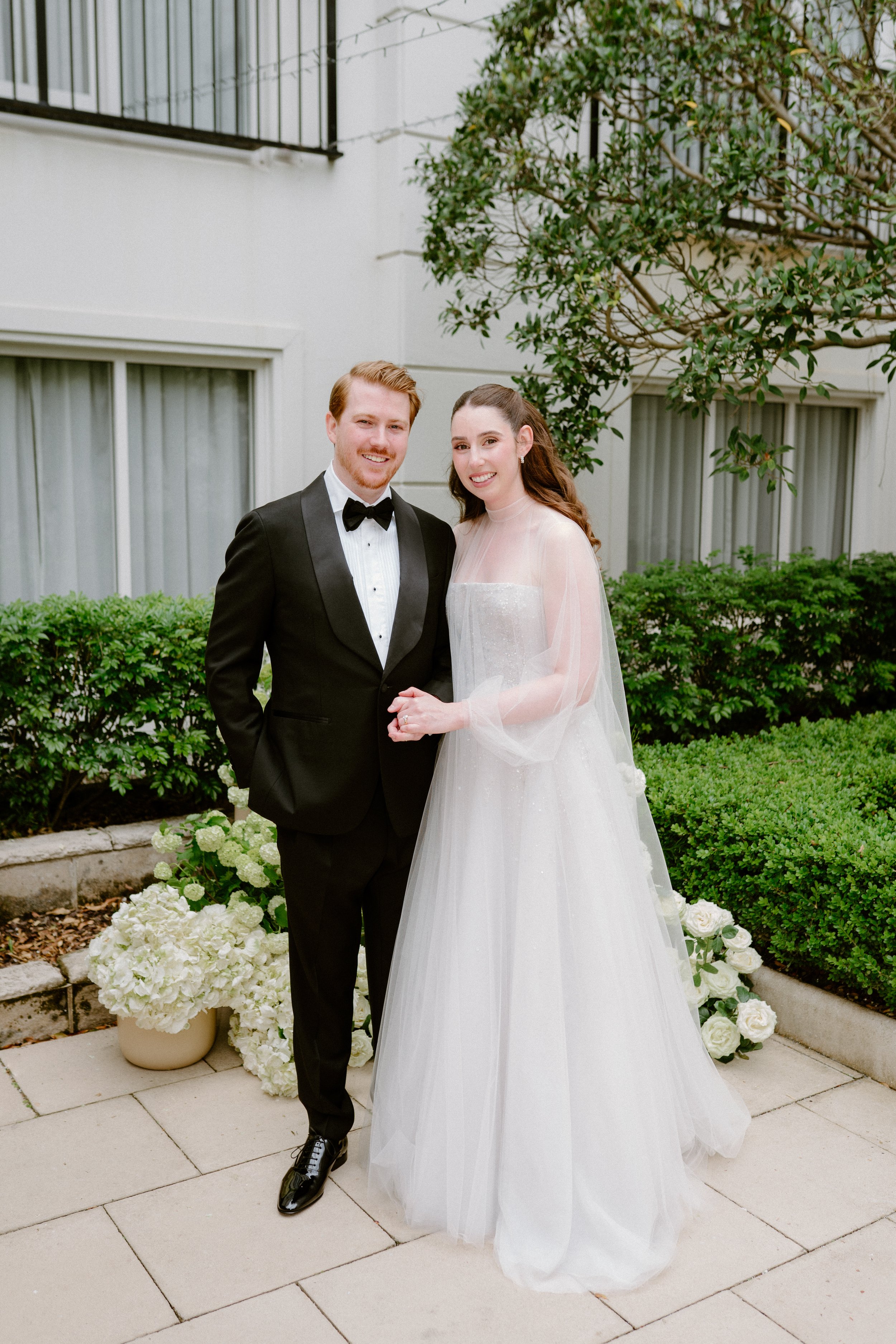 A bride and groom in wedding attire standing outdoors near flowering plants and greenery.