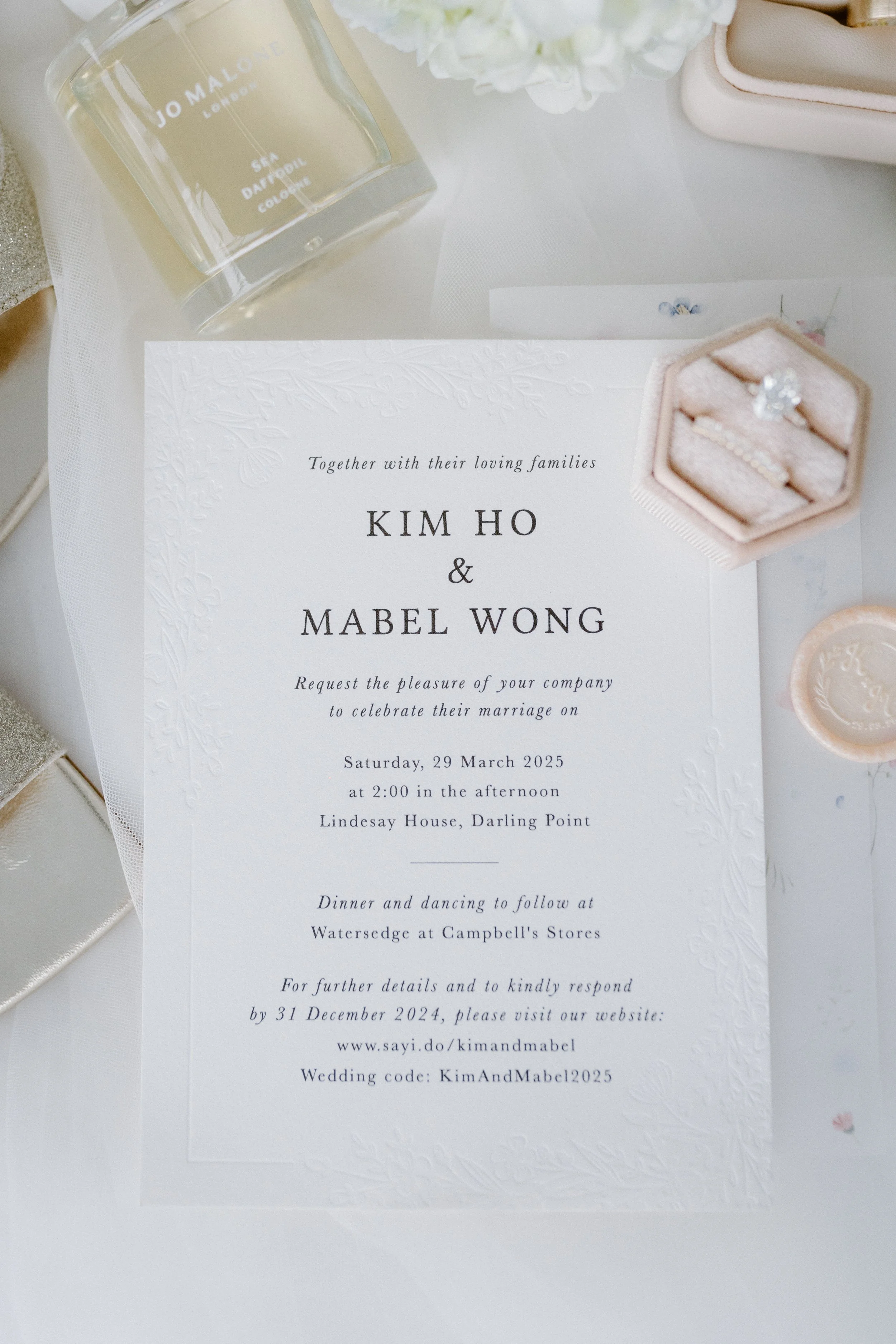 Wedding invitation card with floral embossed border, featuring the names Kim Ho and Mabel Wong, details of the wedding date, time, and location, placed on a white table surrounded by rings in a pink box, a candle, and other decorative items.