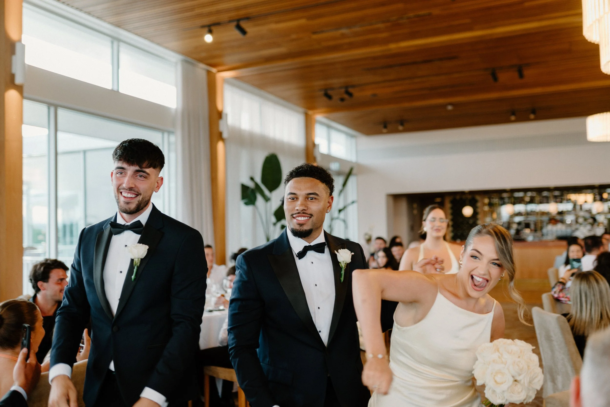 Three people at a wedding reception, two men in tuxedos and a woman in a white dress with a bouquet of white roses, smiling and posing in a decorated hall.
