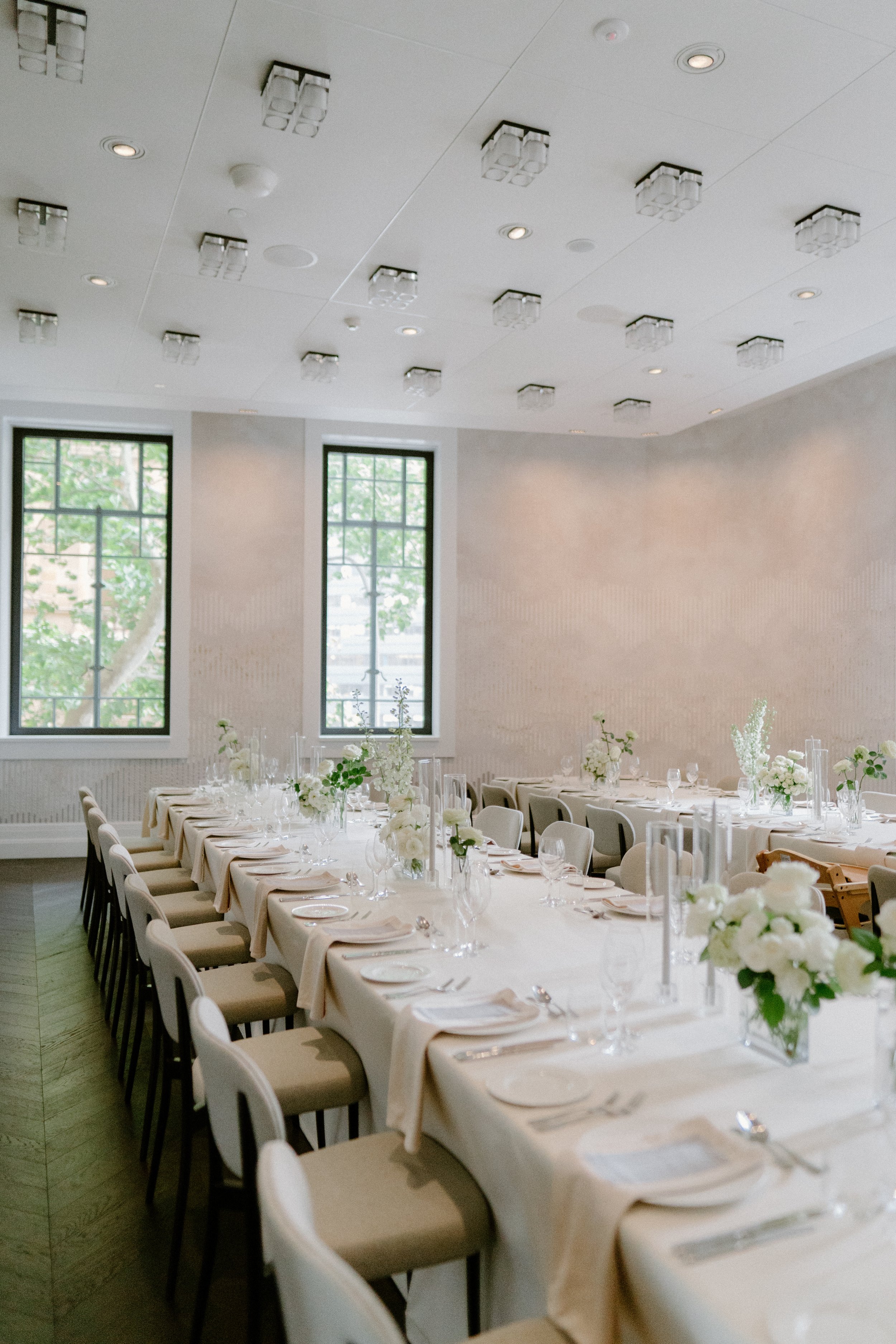 A long rectangular dining table set for a formal event in a bright room with large windows, decorated with white floral centerpieces and elegant tableware.