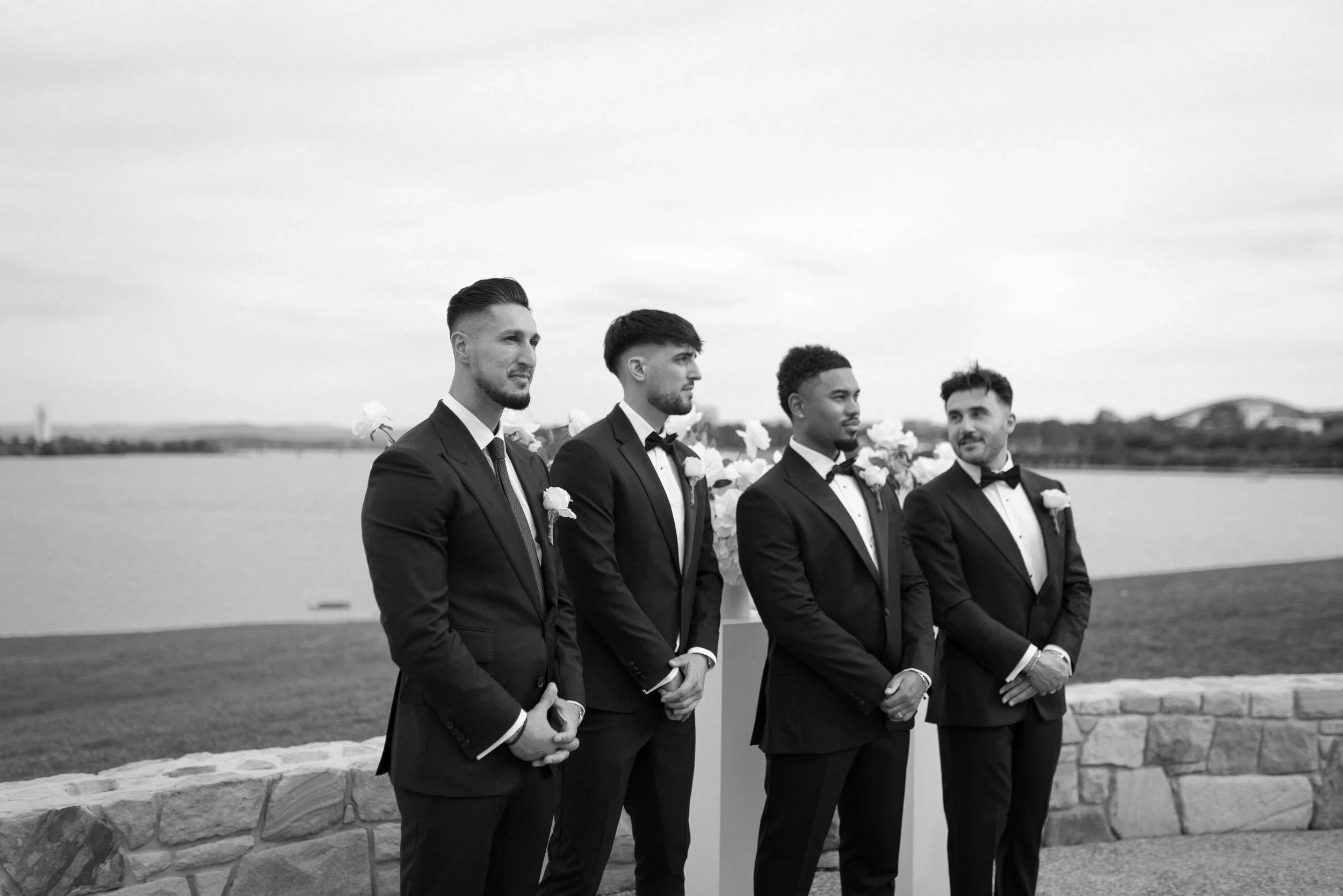 Four men dressed in black tuxedos and bow ties standing outdoors near a body of water, participating in a wedding ceremony.