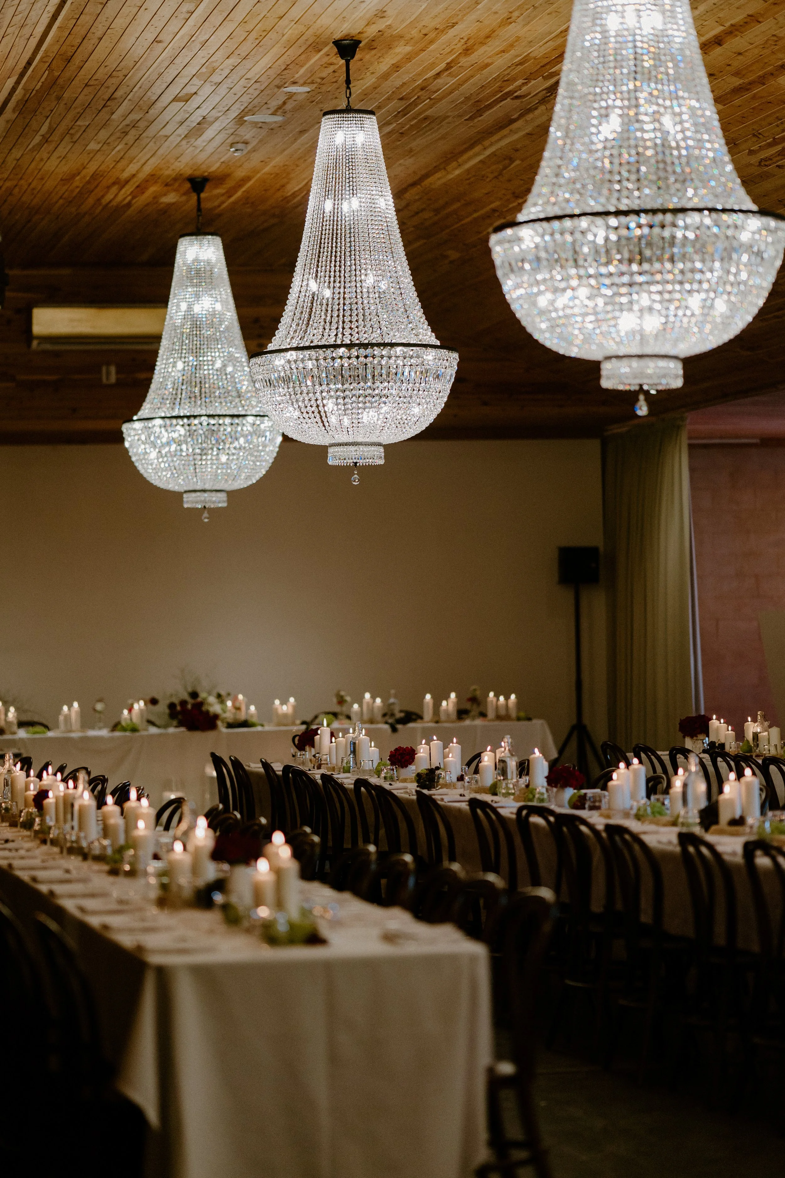 Elegant event space with wooden ceiling, three large crystal chandeliers, and long tables decorated with candles and flowers, set for a gathering or celebration.