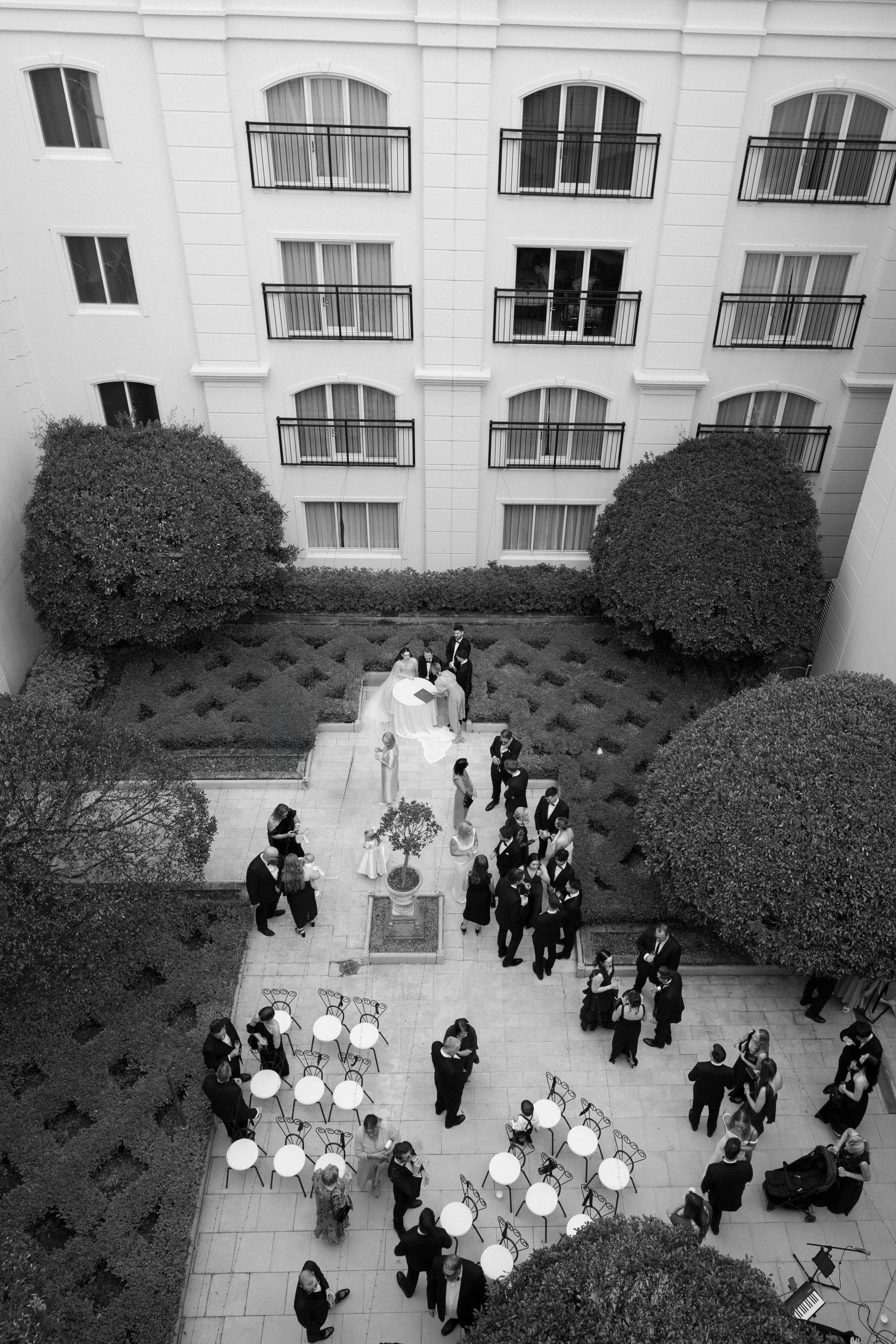 Bird's-eye view of a courtyard with guests dressed in formal attire, wedding ceremony taking place near the top, and tables with chairs set up for gathering.