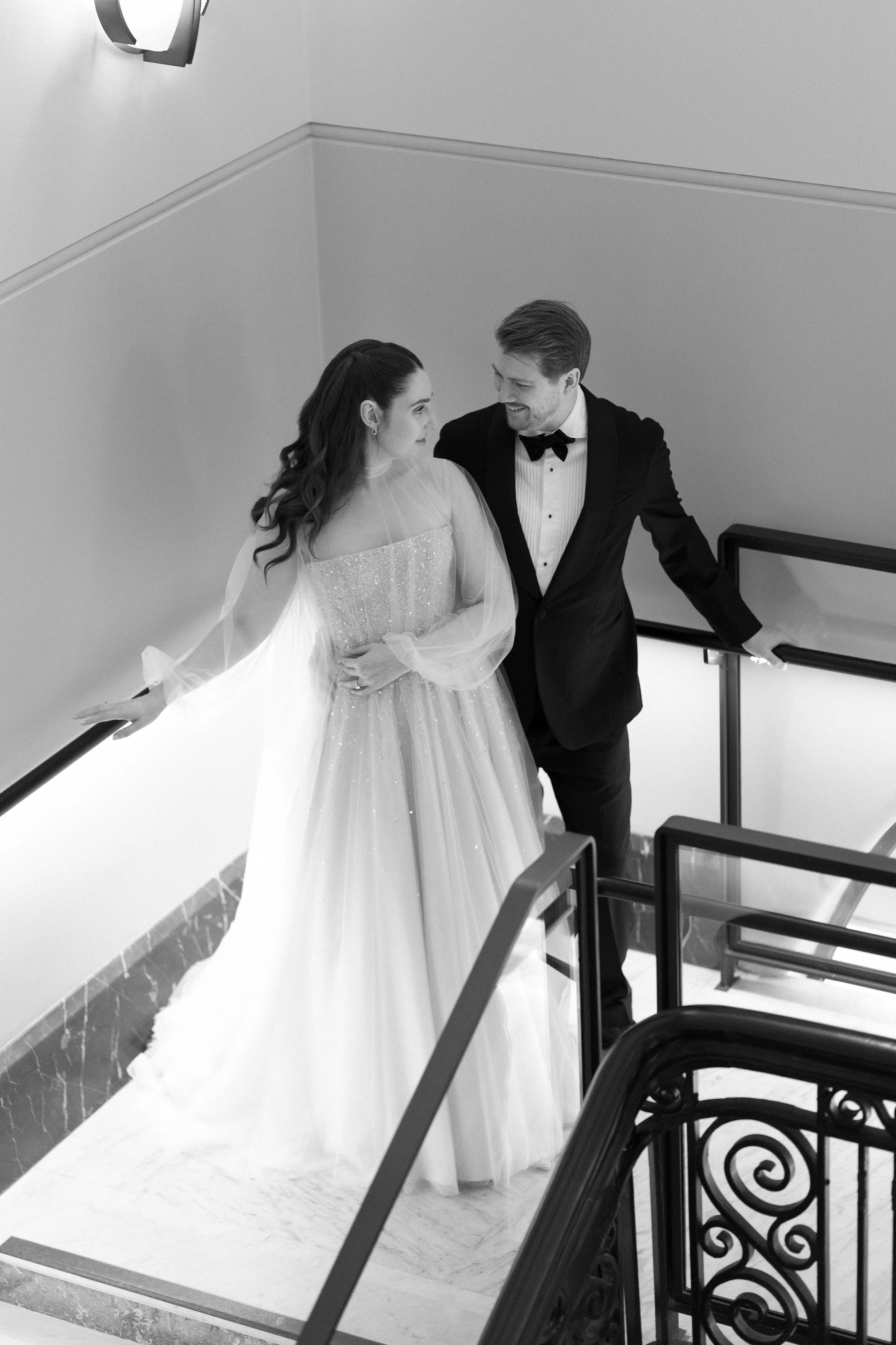 A black and white photo of a bride and groom on a staircase inside a building. The bride is wearing a long, elegant wedding dress with sheer sleeves, and the groom is dressed in a tuxedo with a bow tie. They are looking at each other and smiling.