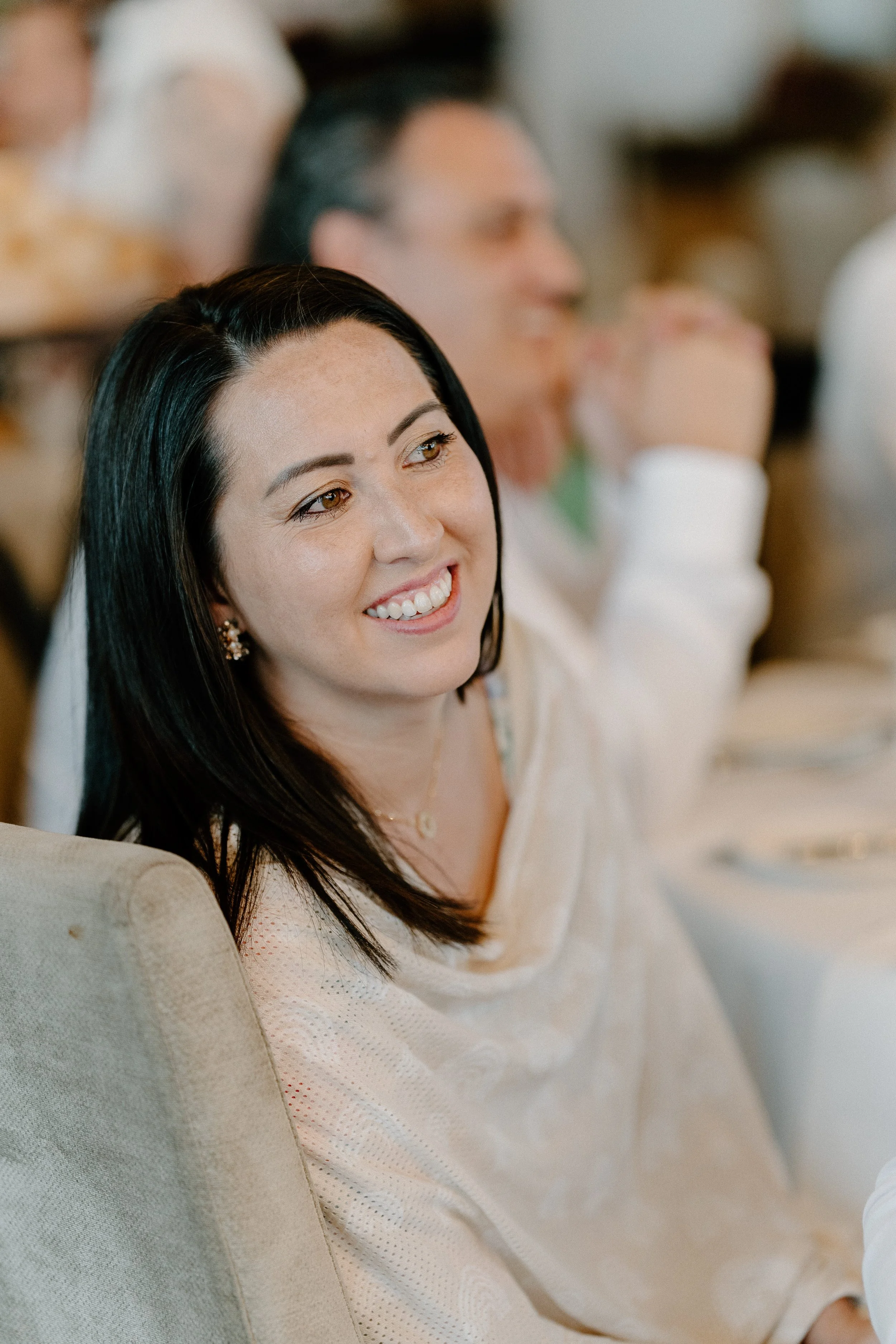 A woman with dark hair and earrings smiling, sitting at a table during an event, with people in the background.