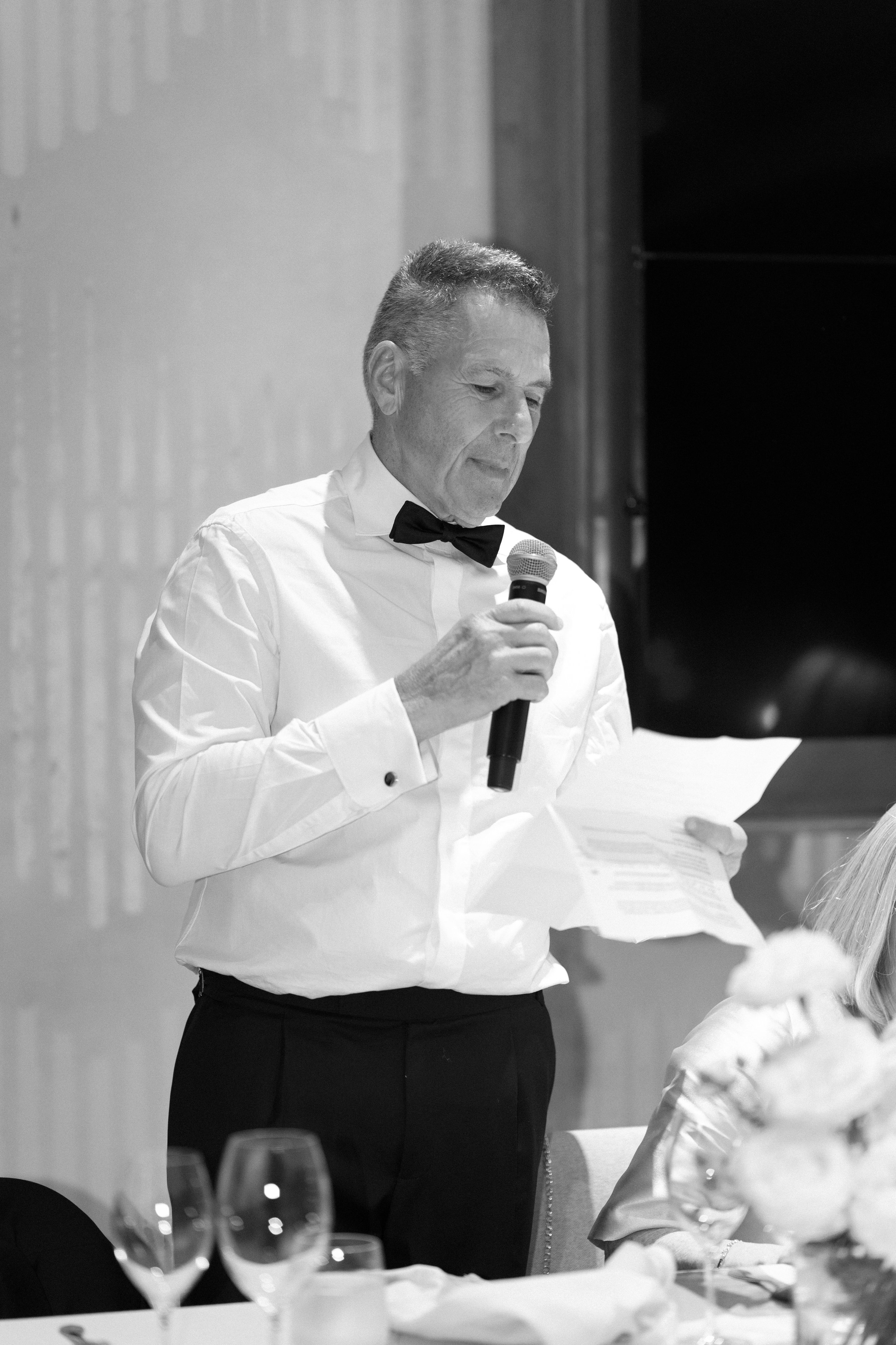 A man in a tuxedo with a bow tie, holding a microphone and reading from a piece of paper at a formal event.