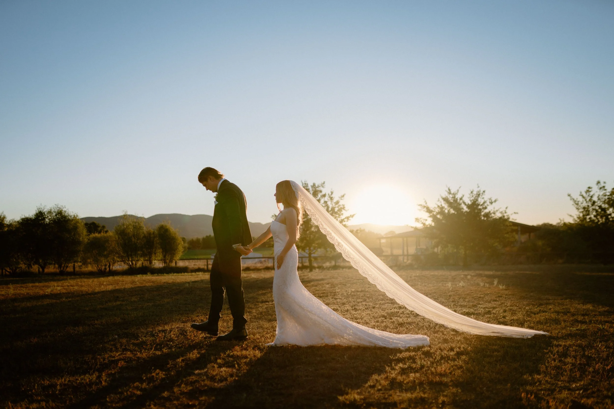 A bride and groom holding hands, walking outdoors during sunset, with the bride's long veil flowing behind her.