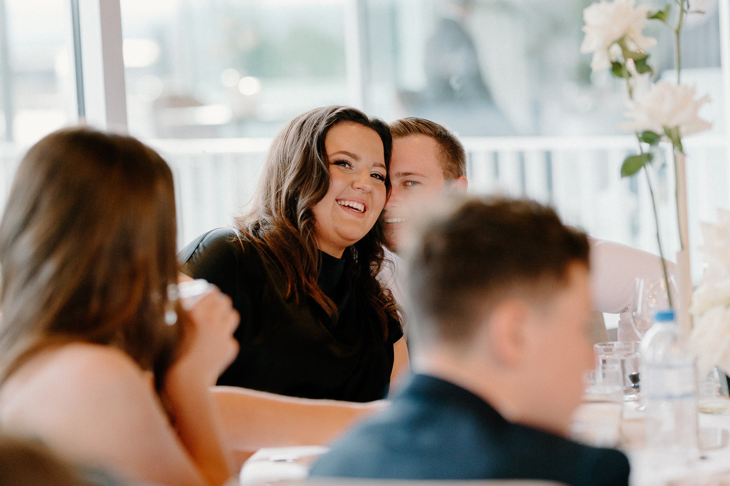 People at a table laughing and talking during a social gathering, with floral decorations in the background.