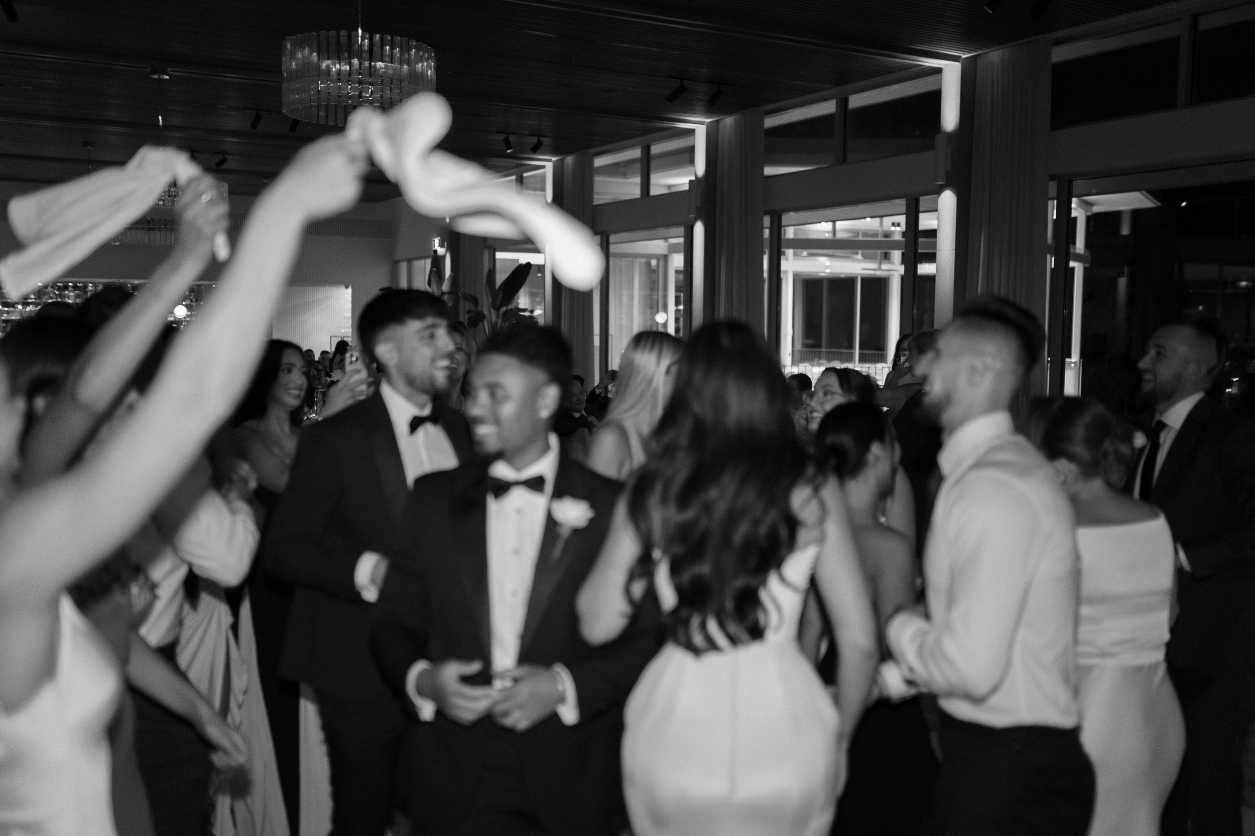 People dancing and celebrating at a formal event in a modern venue with large windows and a chandelier.