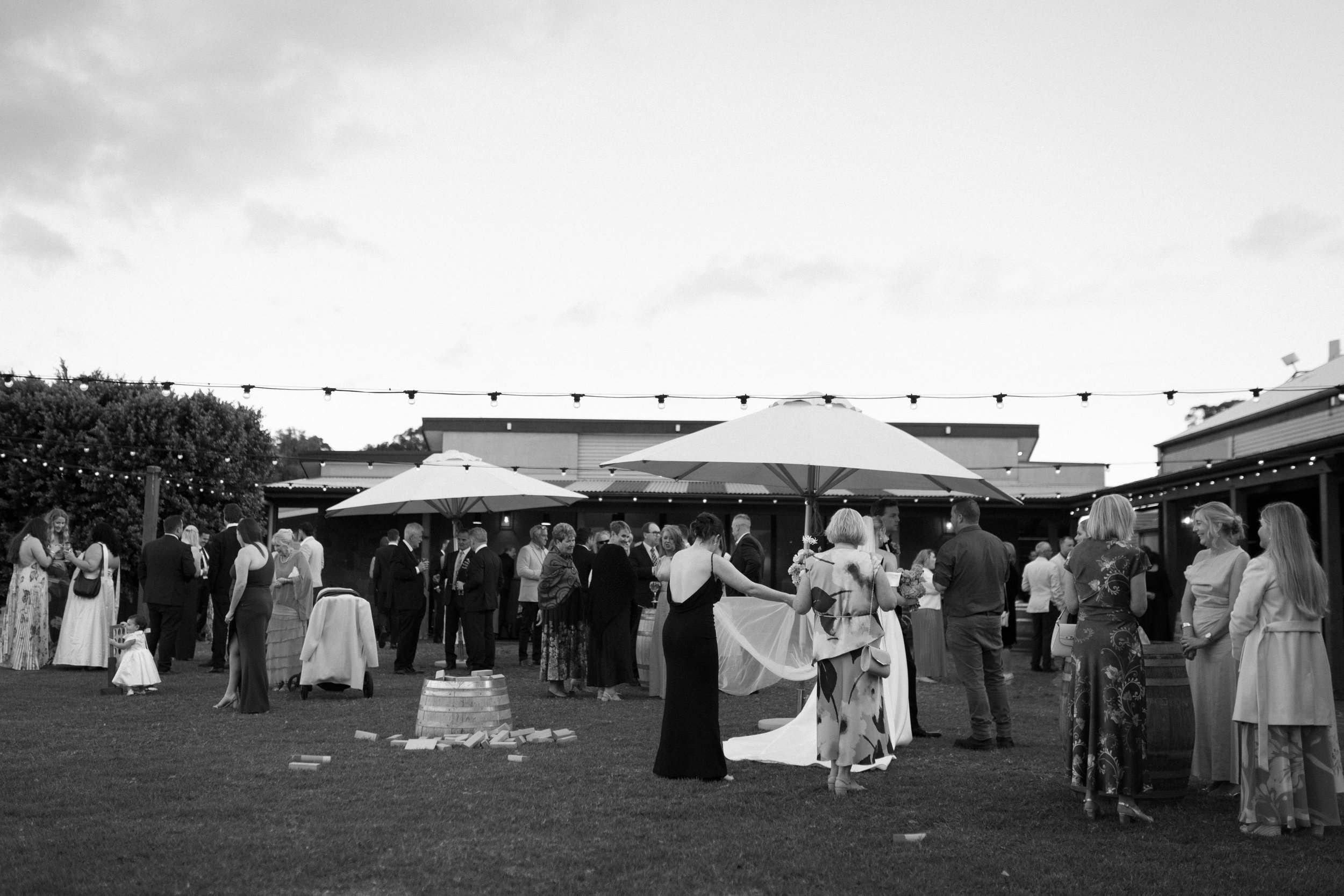 A black and white photo of an outdoor social gathering with people mingling under umbrellas, string lights, and a building in the background.