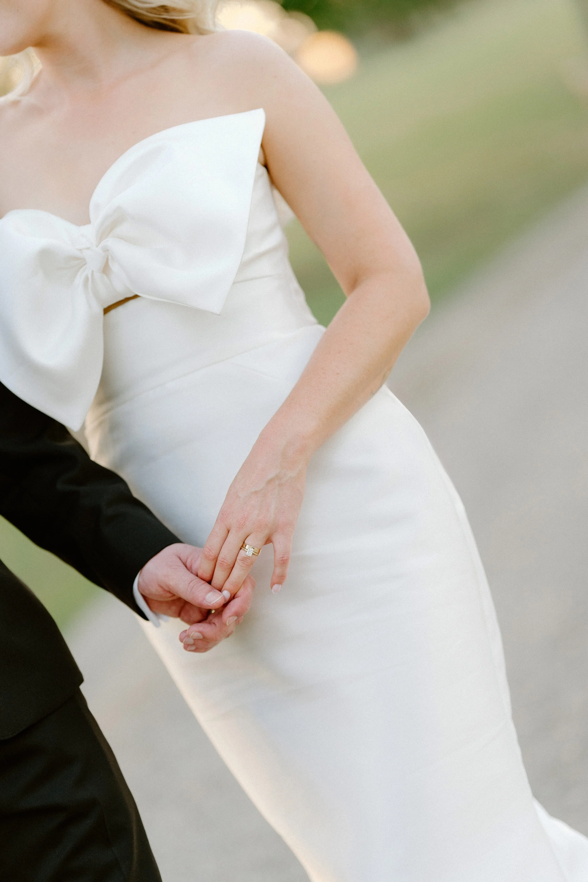 A woman in a white dress with a large bow on the front, holding hands with someone dressed in a black suit. The focus is on the woman and their hands.