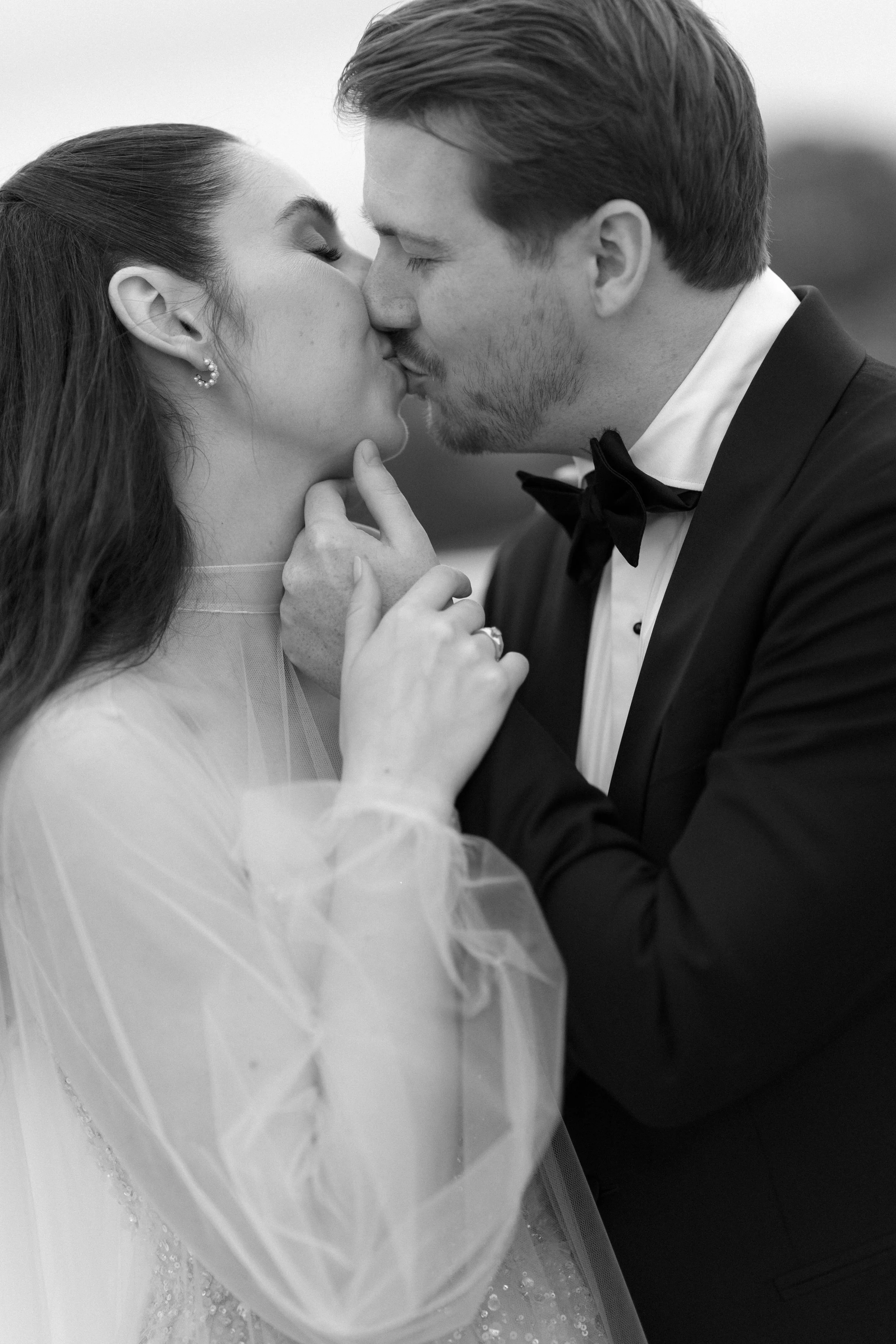 A black-and-white photo of a wedding couple sharing a kiss, with the bride gently touching the groom's chin.