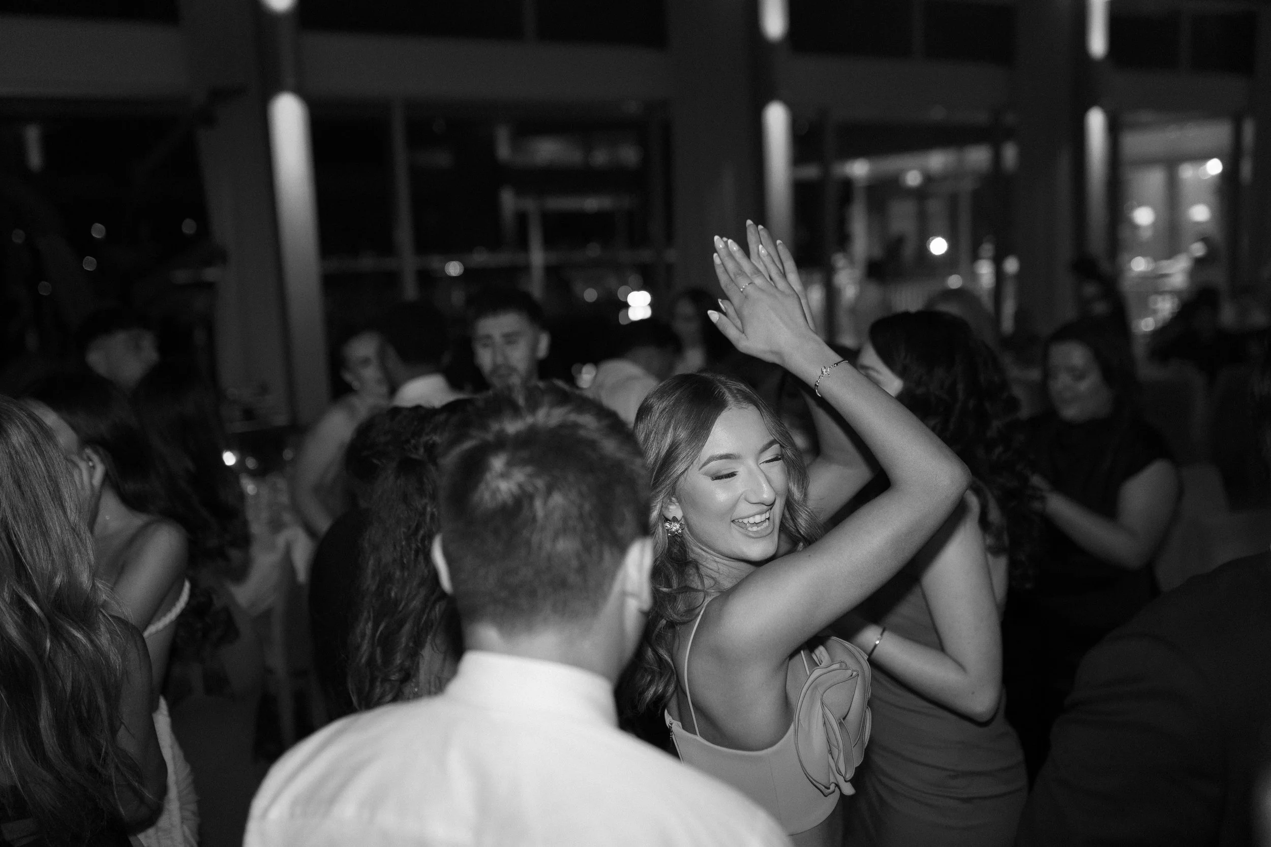 A woman smiling and dancing with her arms raised at a party or celebration, surrounded by other people in a dimly lit indoor setting.