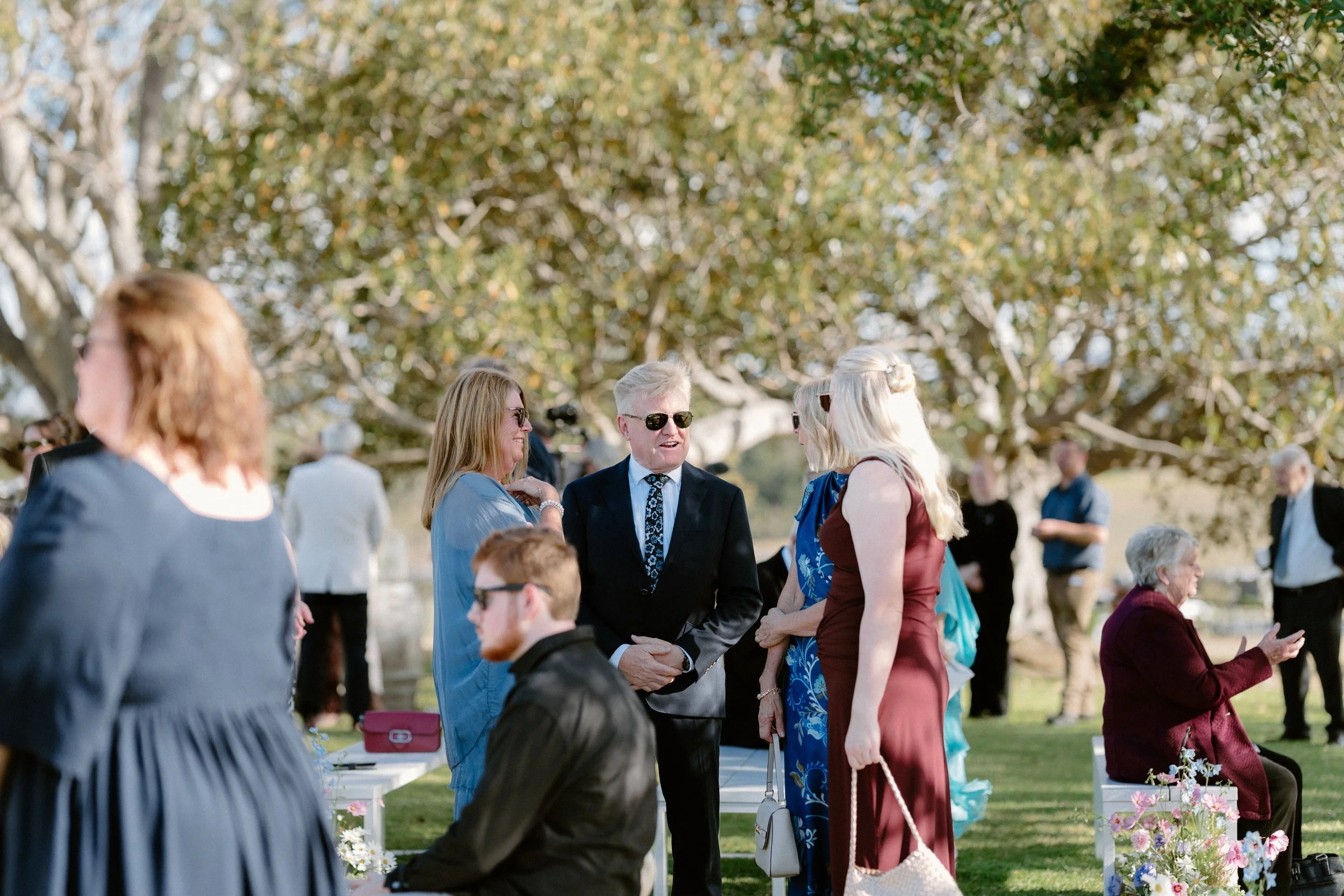 People gathered outdoors for a wedding ceremony, some standing and chatting, with trees in the background.