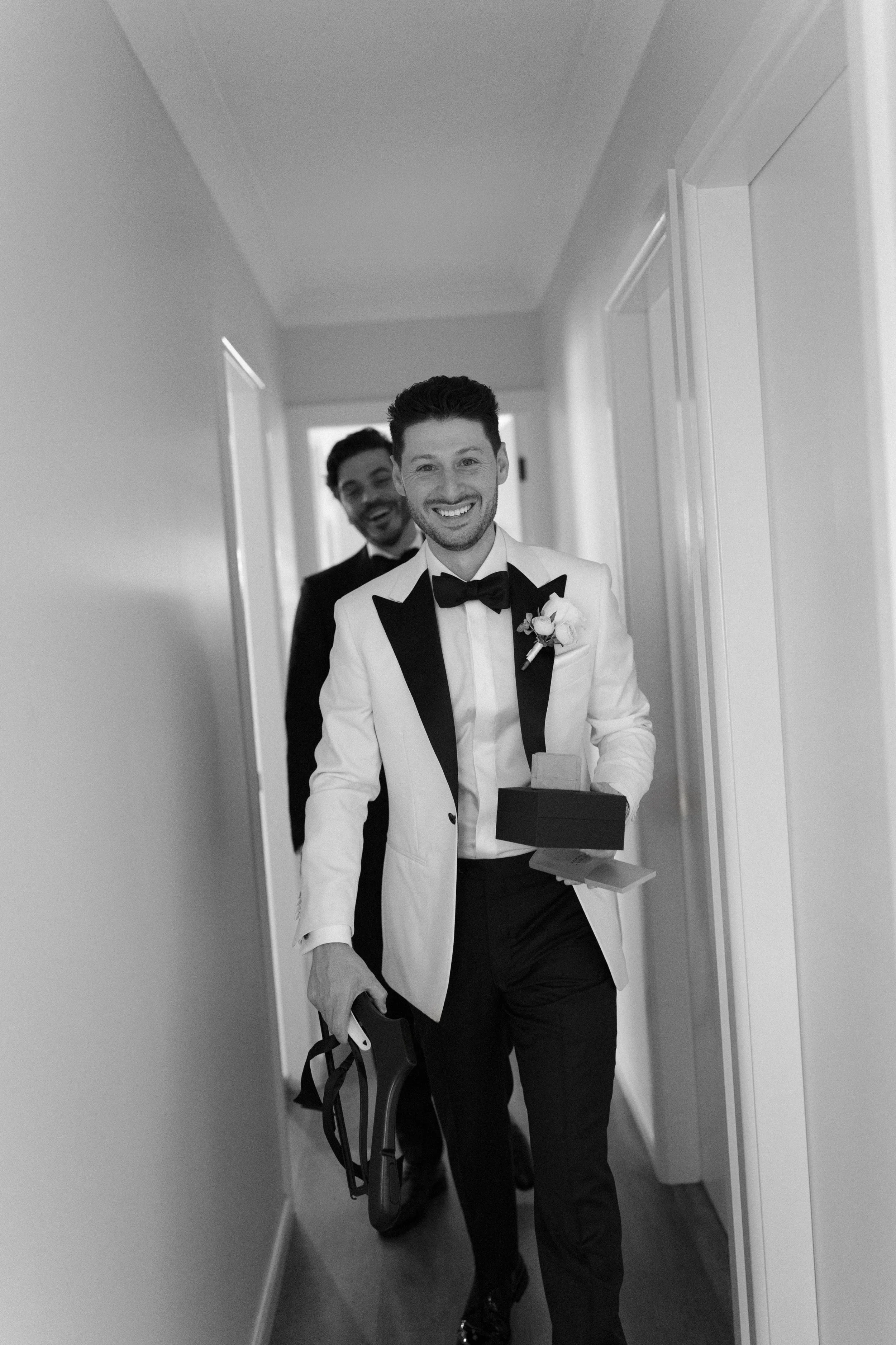 Two men, dressed in tuxedos with bow ties, walking down a hallway; the man in front is holding a gift box and a shoe, smiling, with a boutonniere on his tuxedo's lapel. The man behind is also smiling.