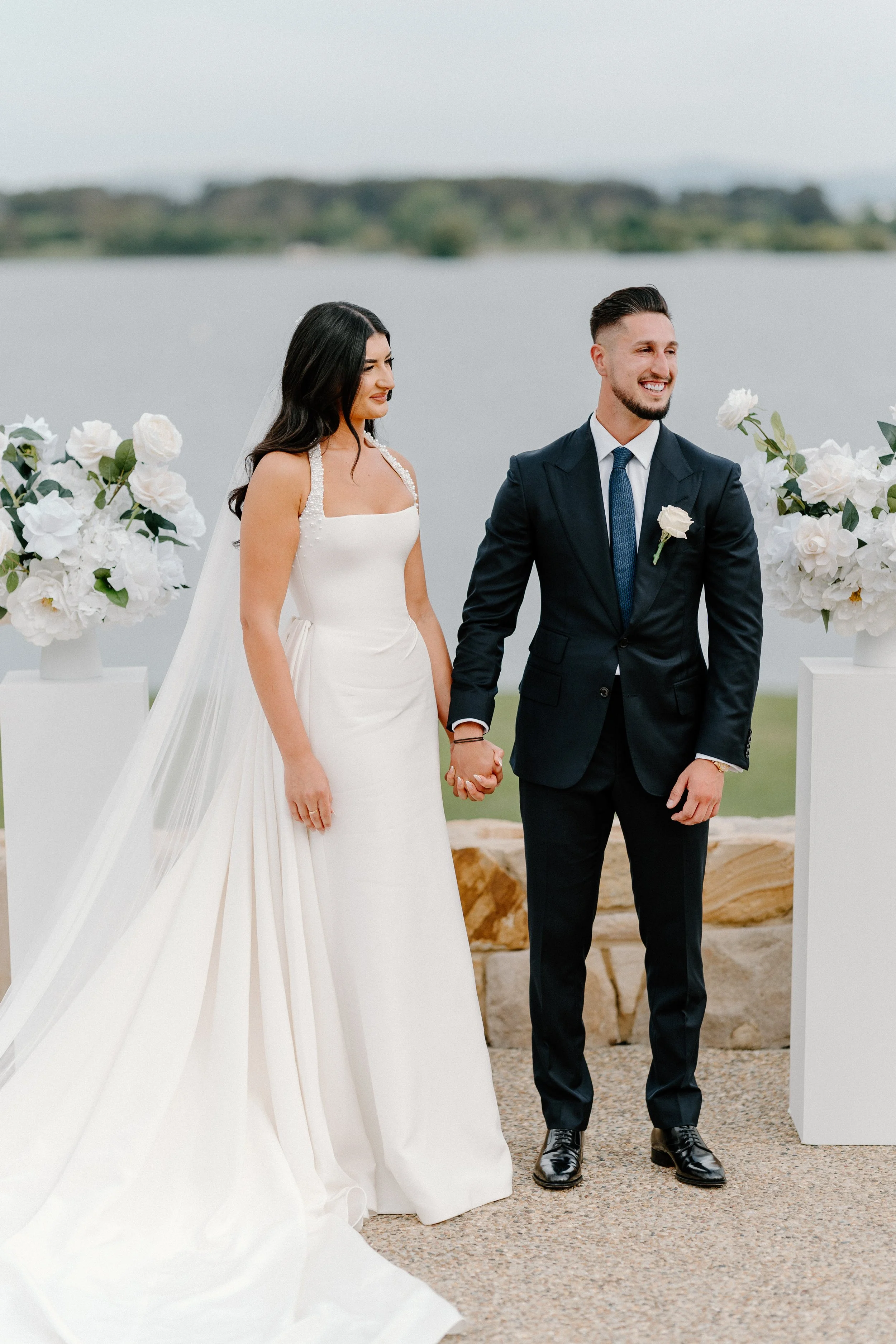 A bride and groom holding hands during their outdoor wedding by the water, with floral arrangements on pedestals behind them.
