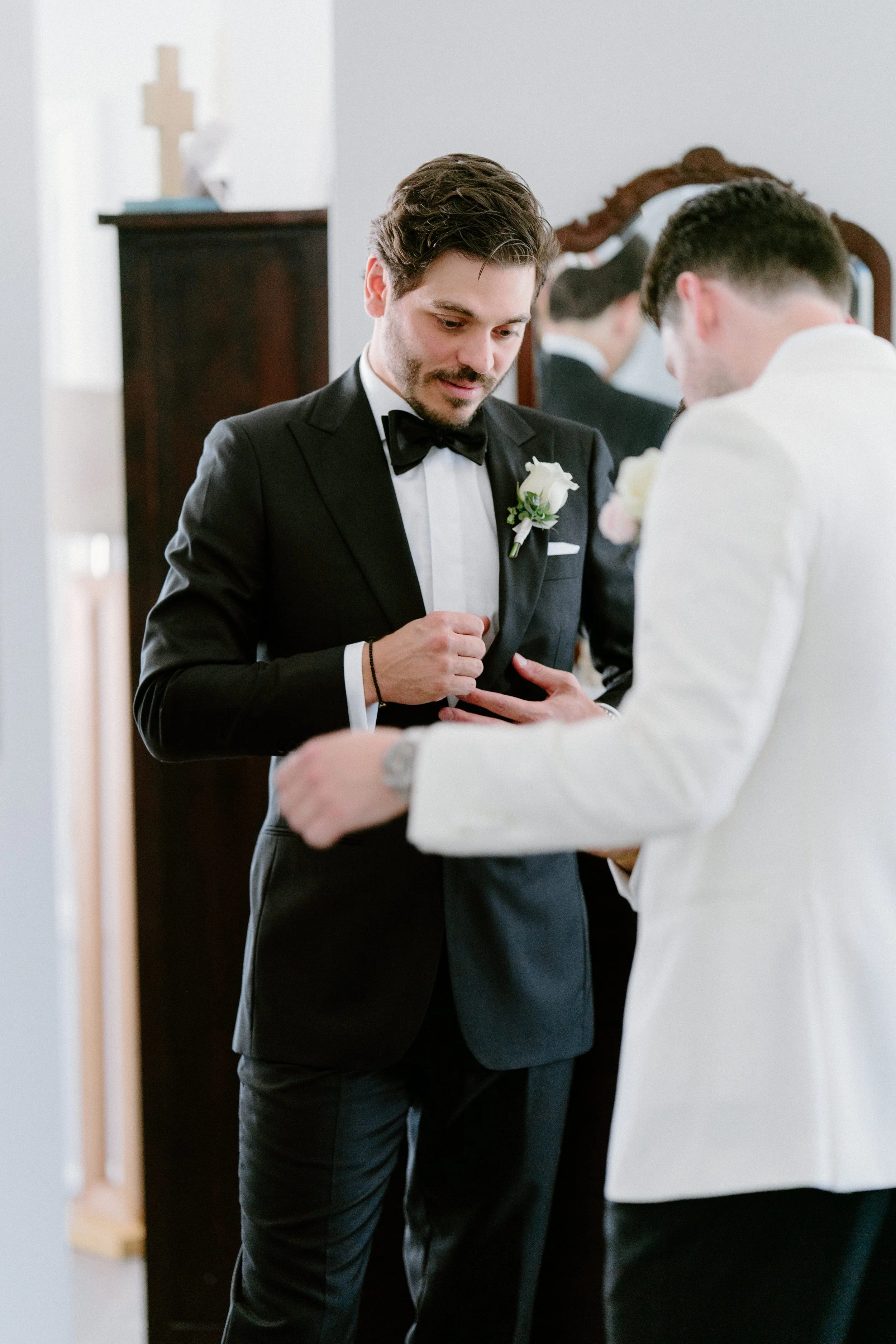 Two men in wedding attire, one in a black tuxedo and the other in a white tuxedo, are sharing a tender moment during a wedding ceremony indoors.