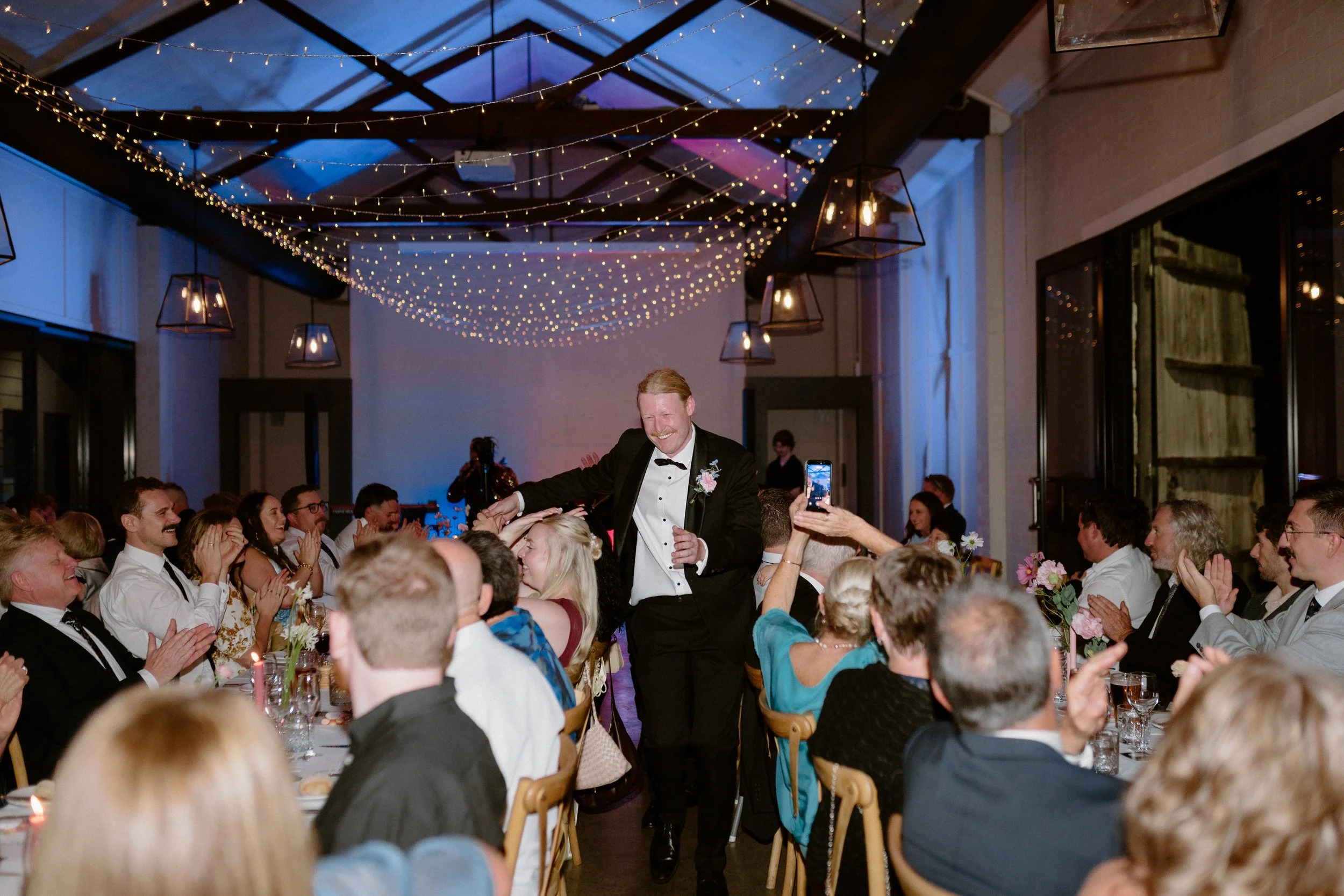 A formal wedding reception with guests sitting at decorated tables, a man in a tuxedo and boutonniere smiling and interacting with guests, and string lights hanging from the ceiling creating a festive atmosphere.