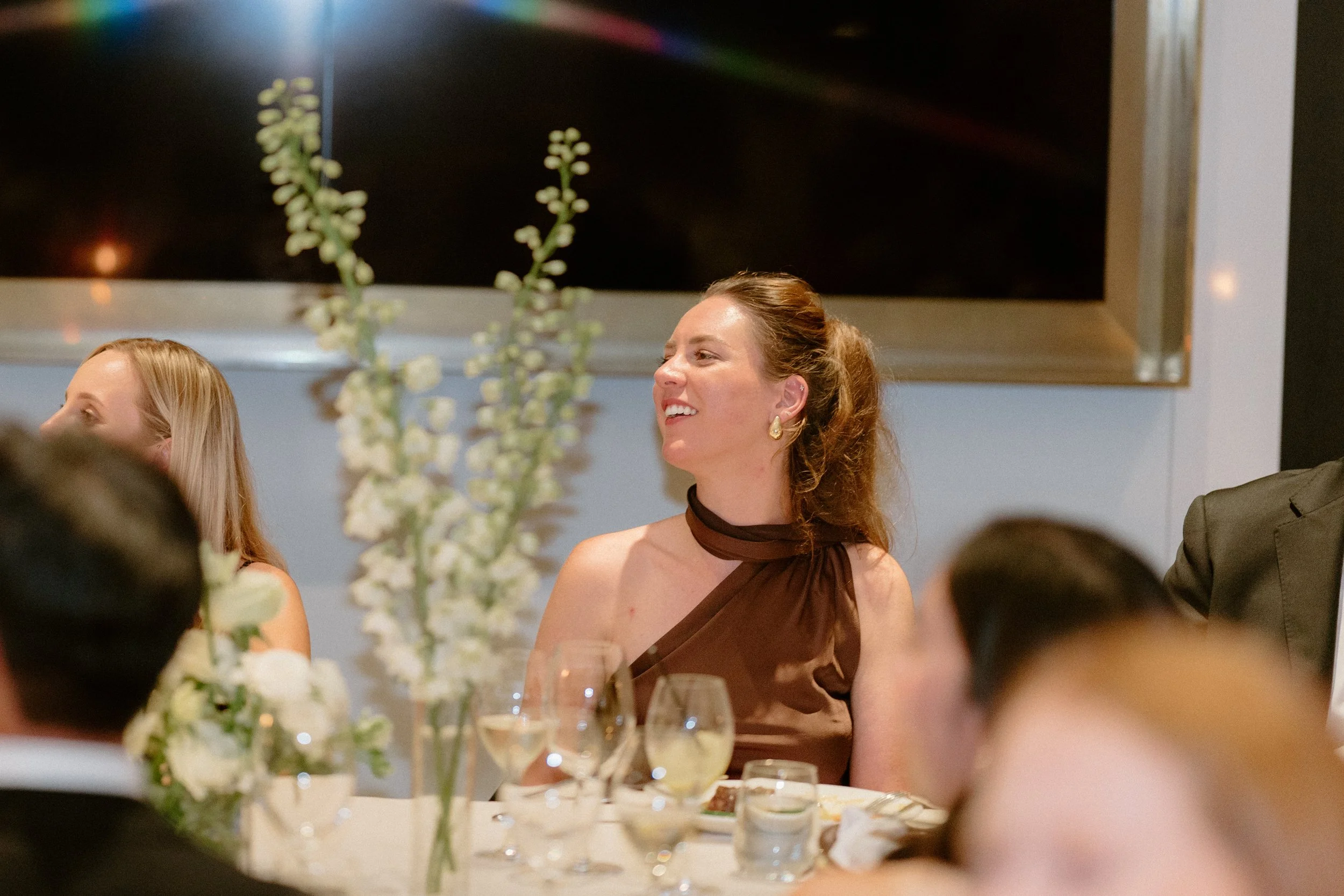 A woman with brown hair in a ponytail, wearing a brown dress with a halter neck, smiling at a formal dinner event with glasses and floral centerpieces on the table.