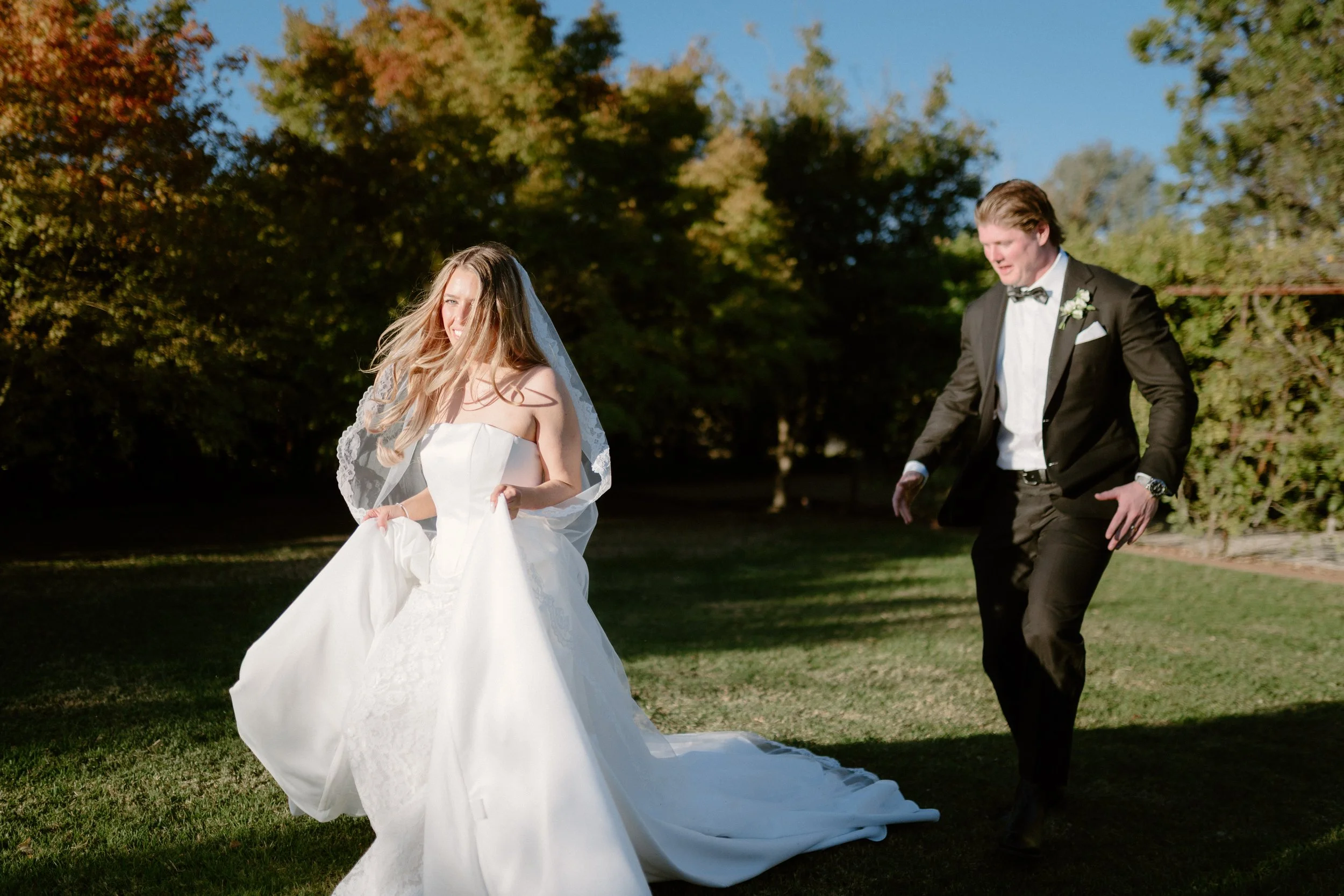 A bride in a white wedding dress with a veil holding her dress, and a groom in a black tuxedo with a bow tie, on a grassy area with trees in the background during daytime.