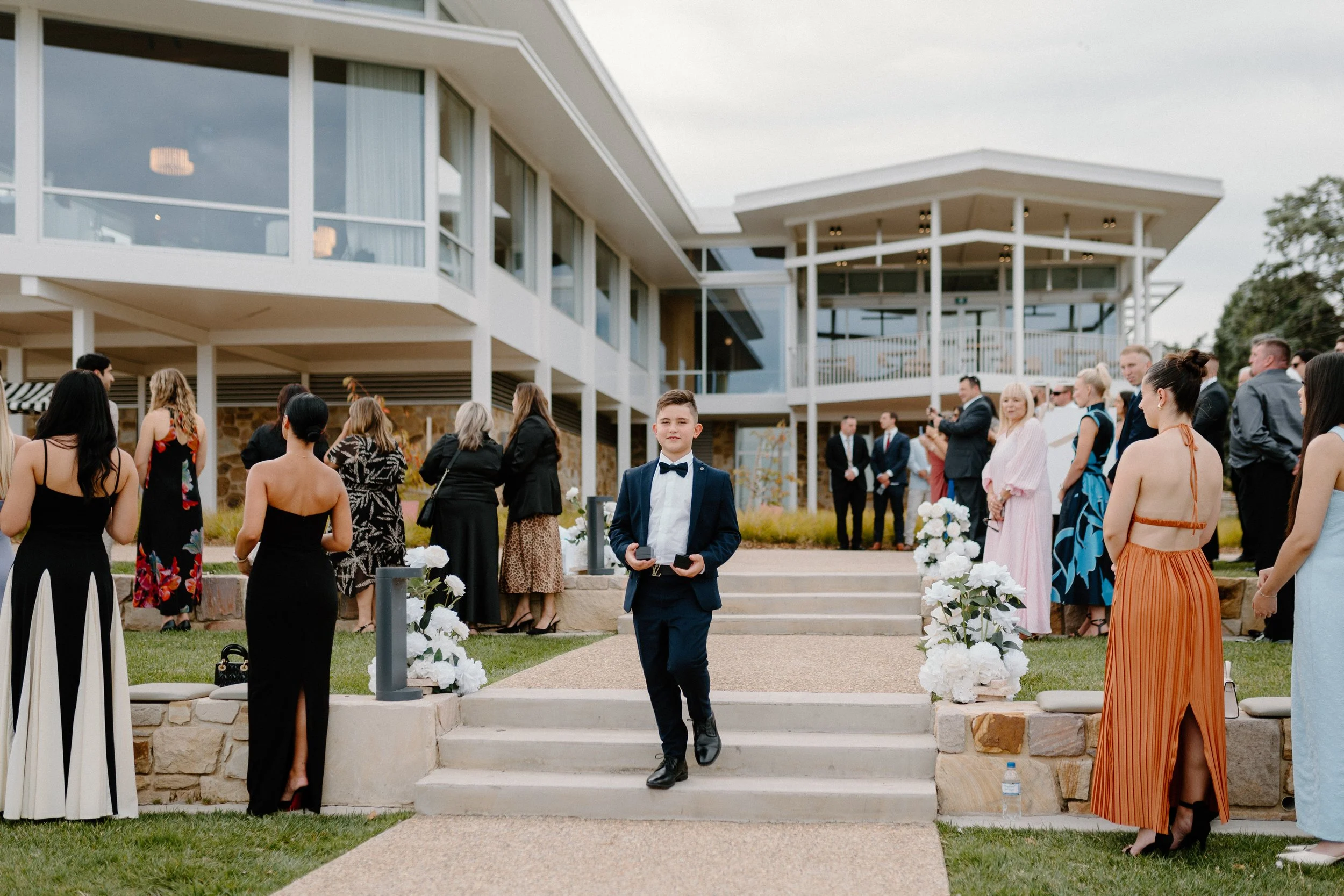 Young man in tuxedo walking down stairs at outdoor formal event with guests standing on either side, large modern building in background, decorated with white flowers.