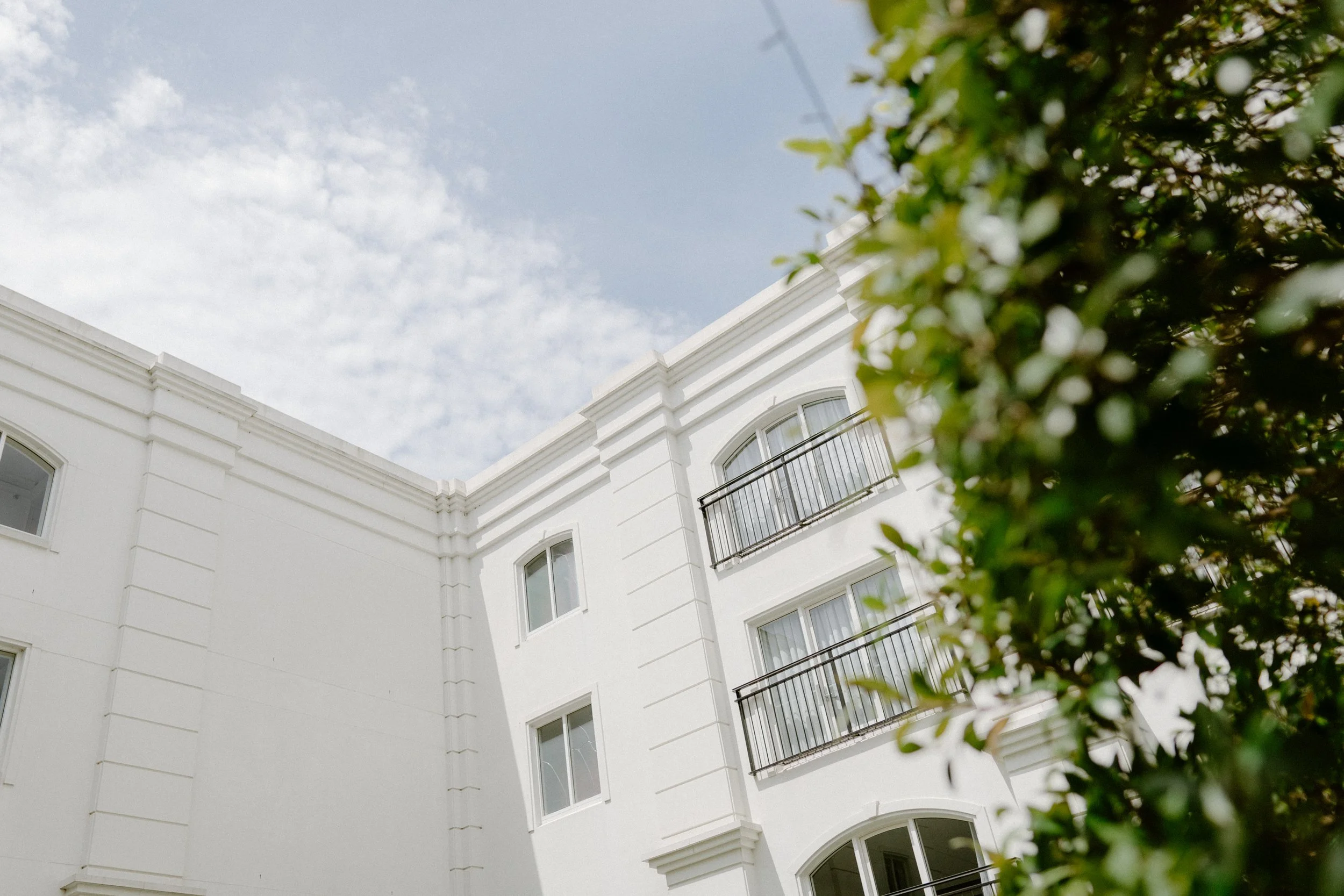 White multi-story building with balconies and windows, partly obscured by green foliage, against a cloudy sky.
