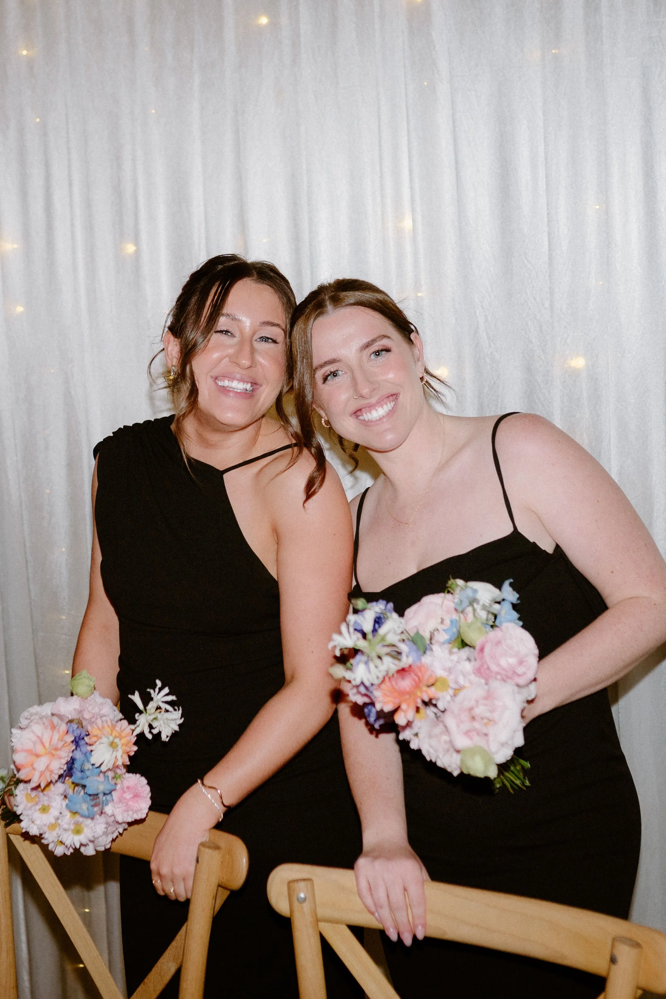 Two women wearing black dresses and holding bouquets of pink, blue, and white flowers, smiling and posing indoors against a white curtain with lights.