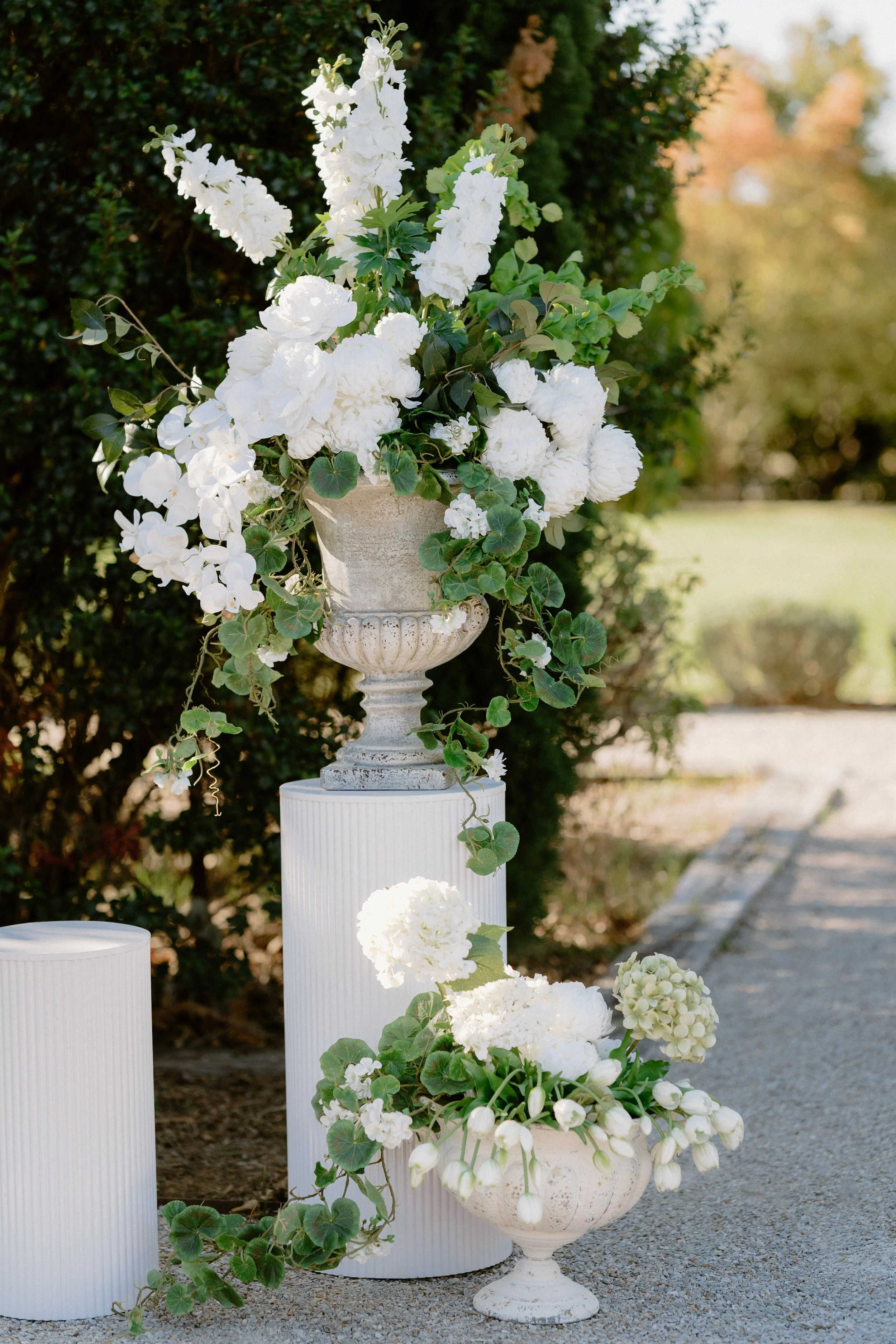 A floral arrangement of white flowers in decorative urns outdoors in a garden setting, with greenery and a pathway in the background.