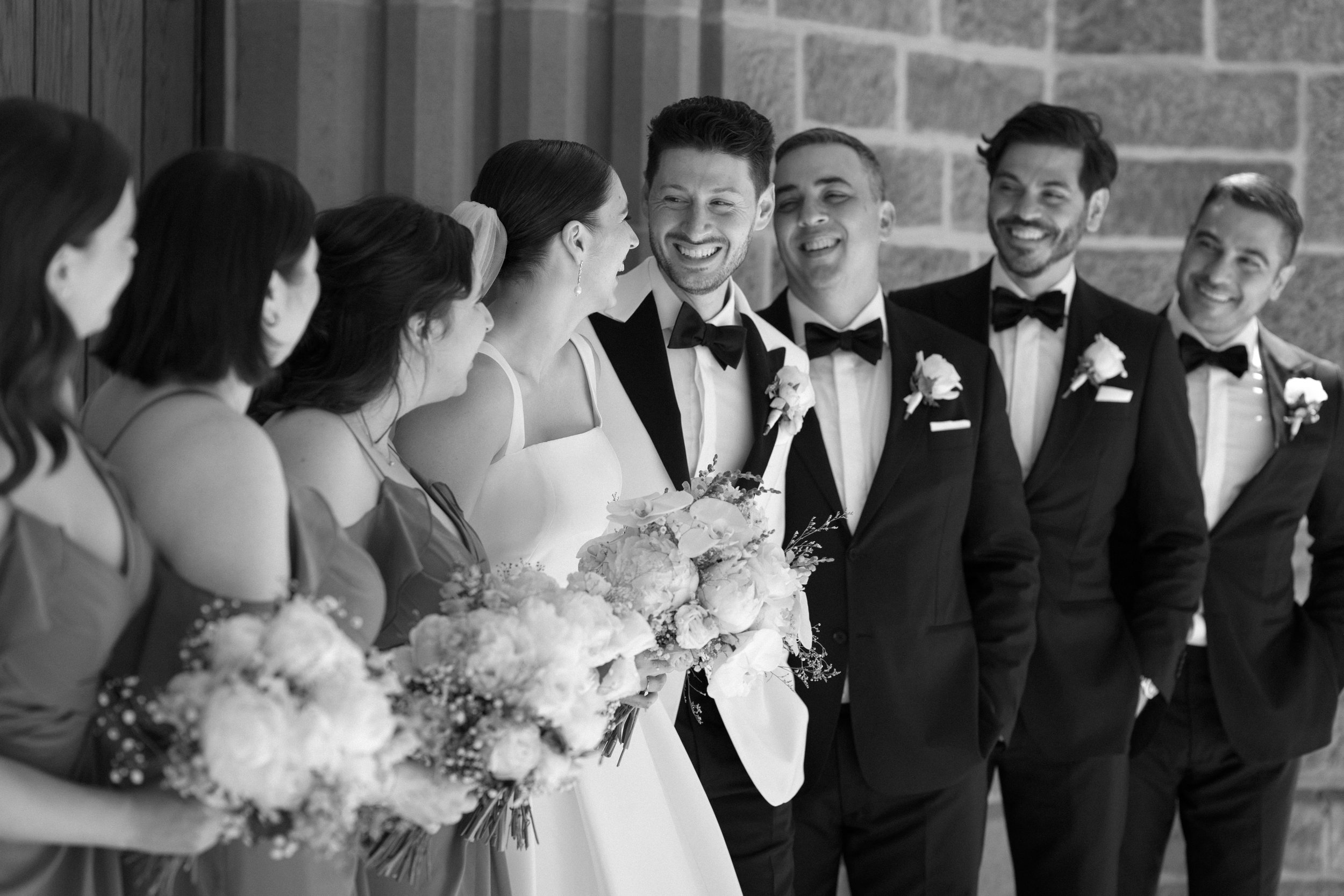 A black and white photo of a bride and groom with their wedding party. The bride is holding a bouquet and smiling, surrounded by bridesmaids and groomsmen in formal attire, all smiling and looking at each other.