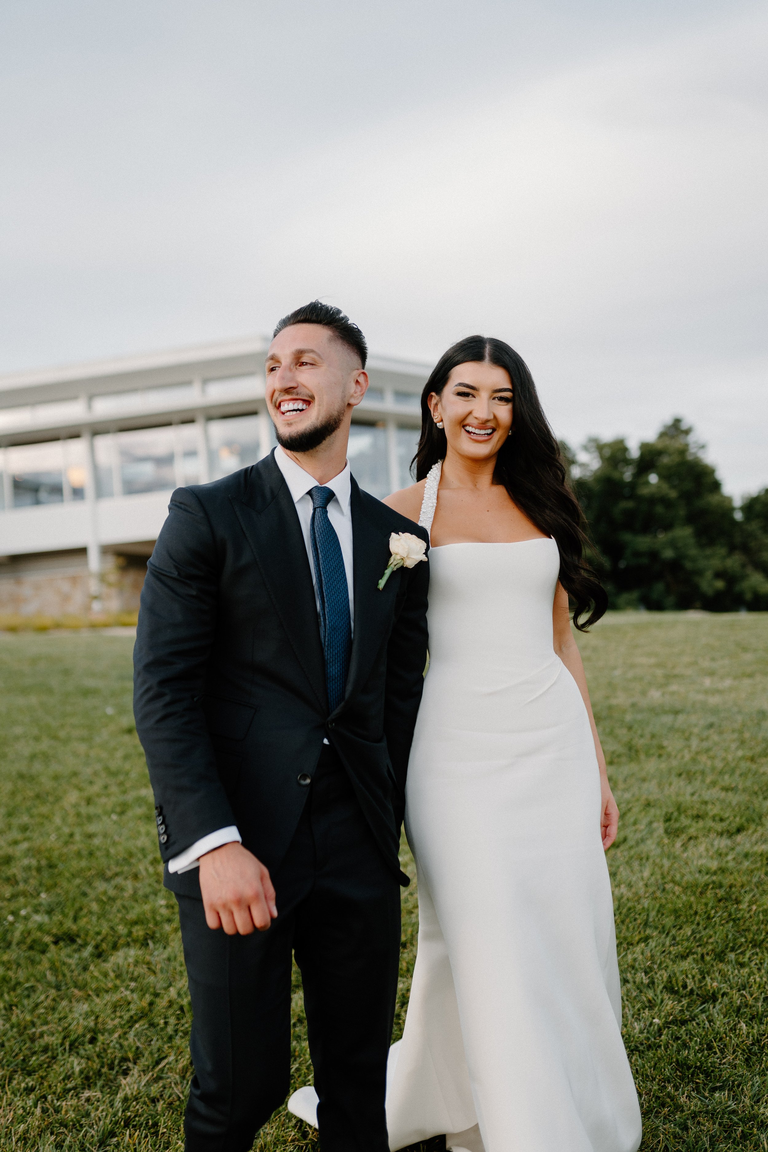 A newlywed couple standing on a grassy field in wedding attire, smiling and enjoying a moment together outdoors, with a modern building in the background.