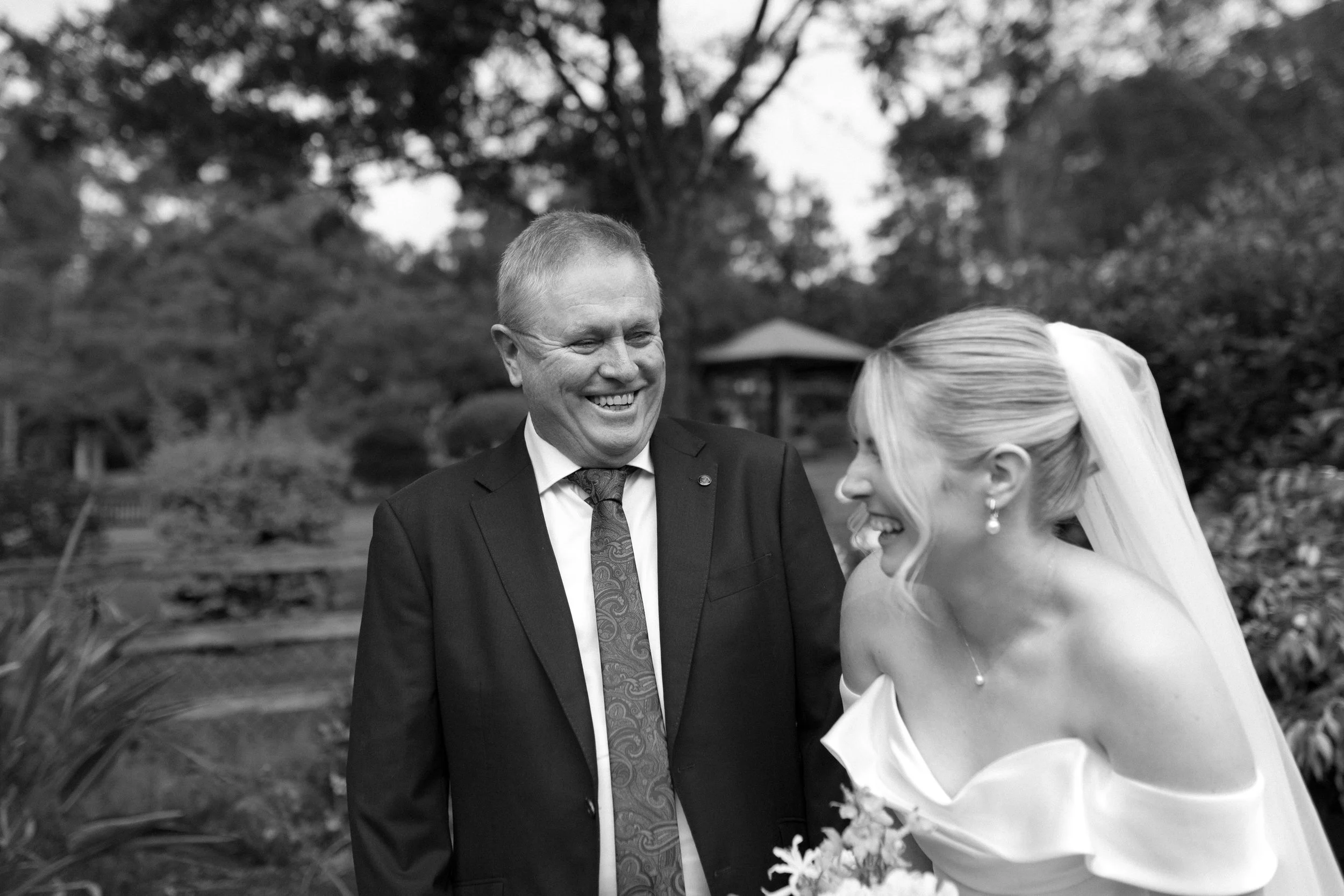 A smiling man in a suit and tie looking at a woman in a white dress with a veil, outdoors with trees and a gazebo in the background.